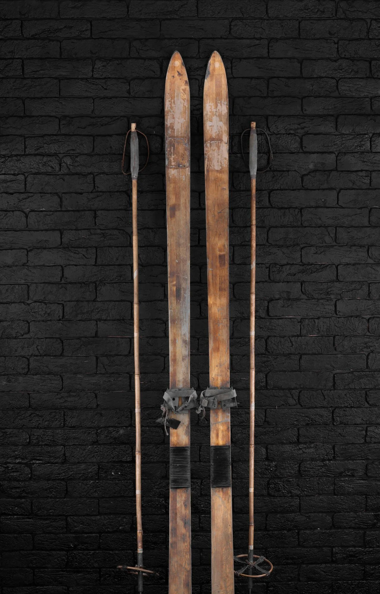 Vintage wooden skis with leather straps and ski poles against a black brick wall