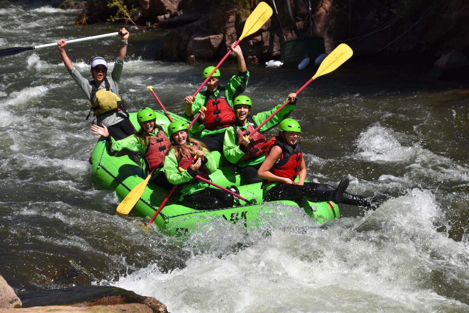 Group of smiling people wearing helmets and life jackets white water rafting in a river.