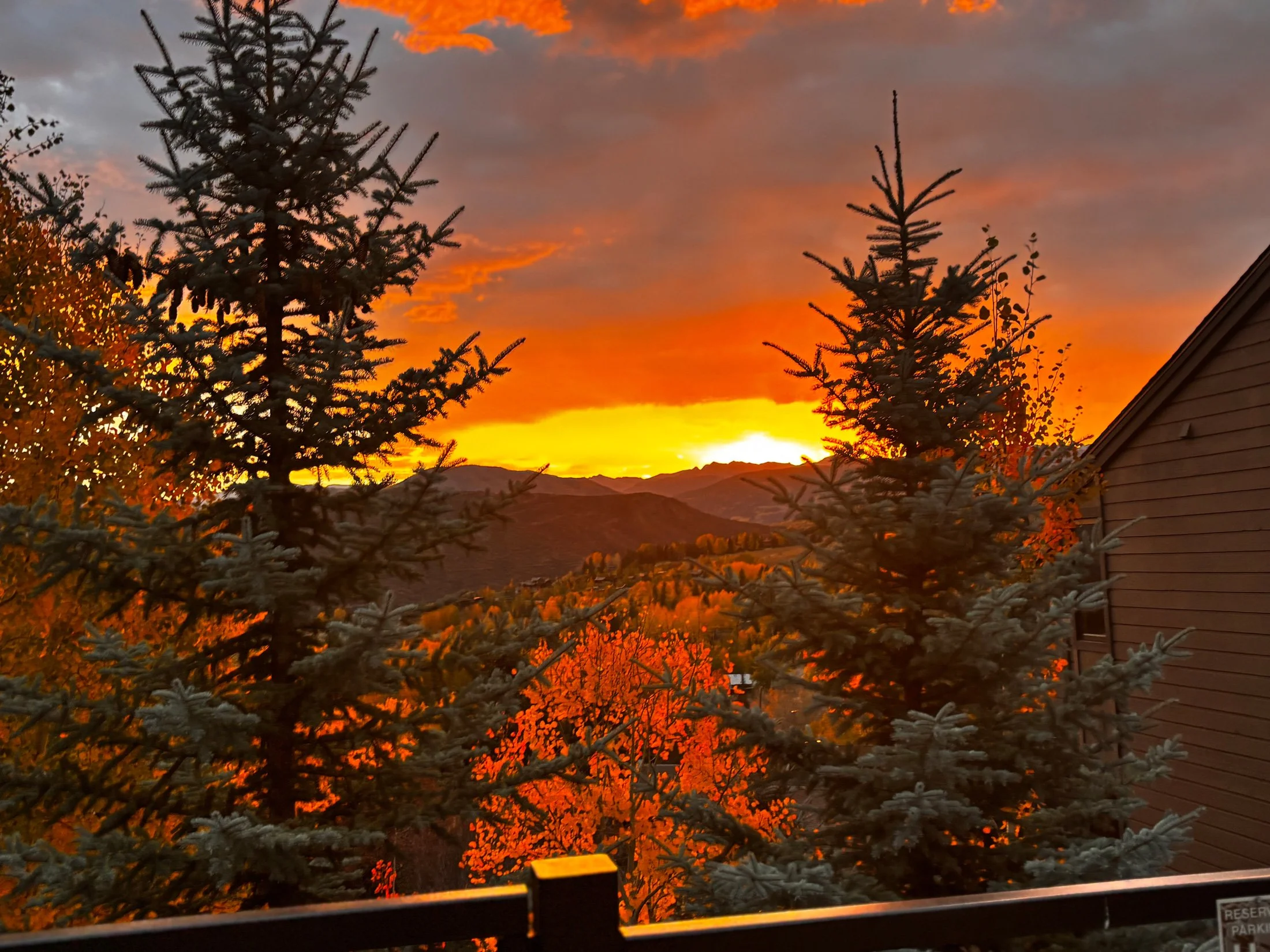 A vibrant sunset over mountains, with orange, yellow, and purple hues in the sky, framed by evergreen trees and a brown house on the right.