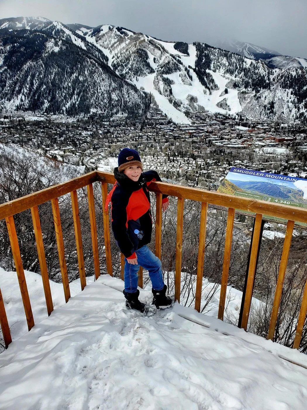 A young boy in winter clothing standing on a snowy observation deck with a mountainous, snow-covered landscape and a small town in the valley behind him.