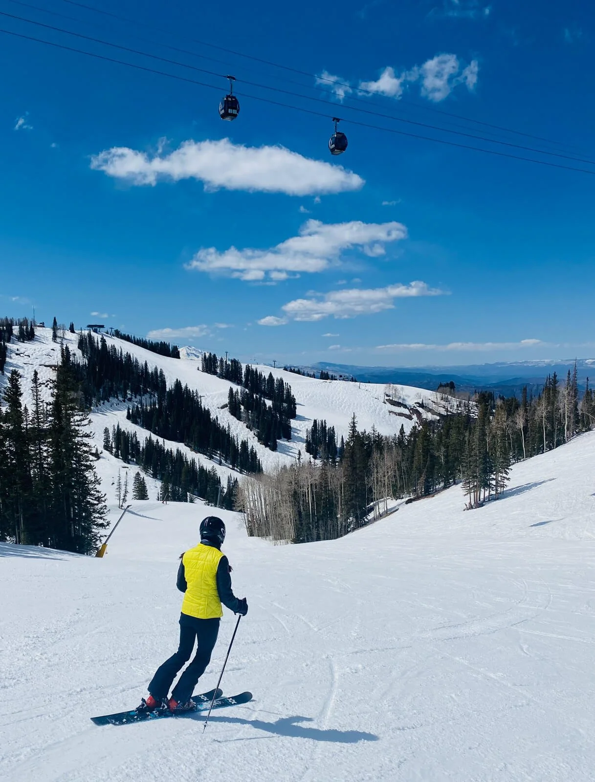 A skier wearing a yellow vest and black helmet skiing on a snowy mountain slope with trees and ski lifts in the background, under a blue sky with clouds.