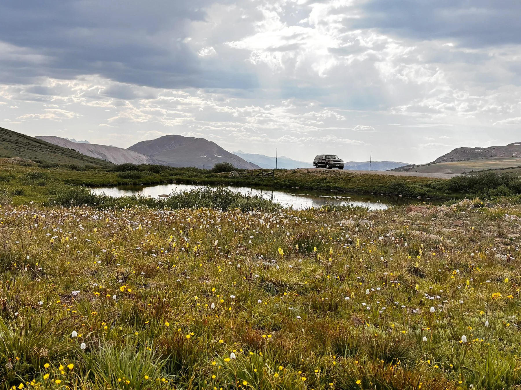 Scenic landscape with a small pond, wildflowers in the foreground, rolling hills, mountains in the background, and a cloudy sky with sunlight breaking through.