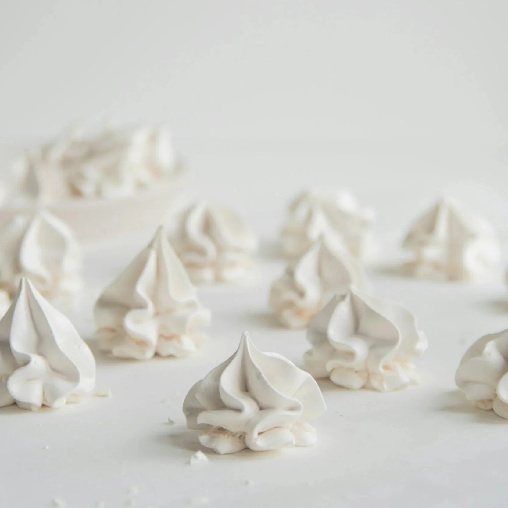 Multiple white meringue cookies scattered on a white surface with a white bowl in the background.