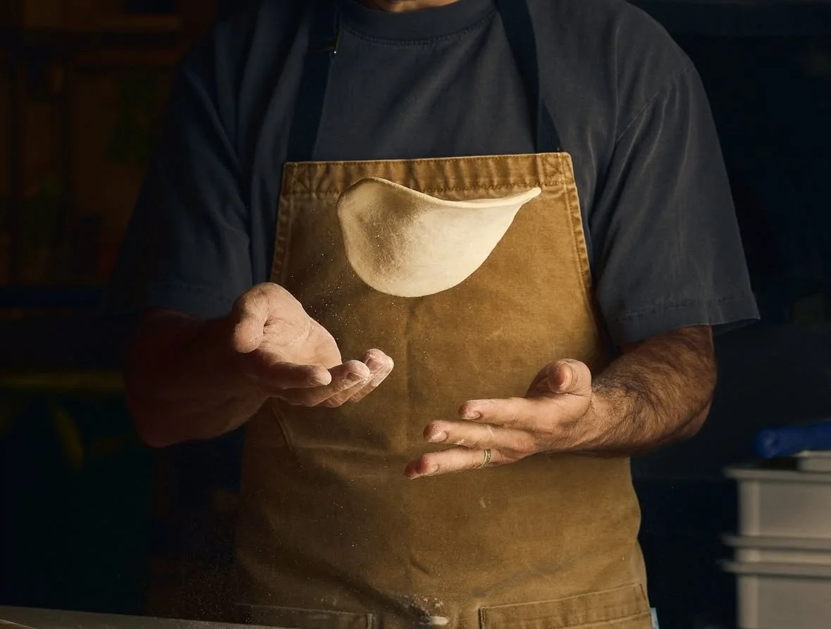 A person wearing a dark shirt and an apron, holding a handful of flour with a piece of dough suspended above the flour in the air.