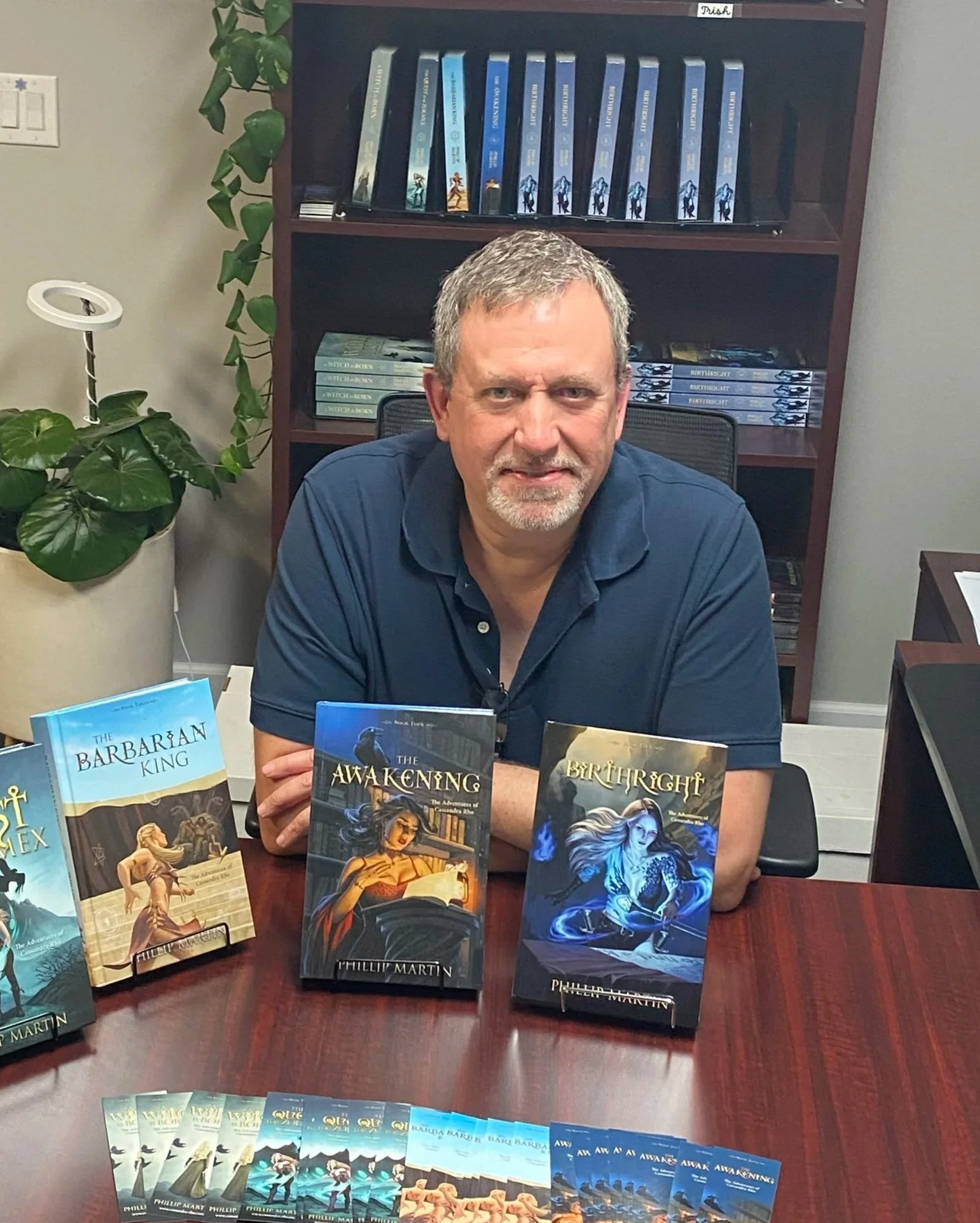 A man with gray hair and a beard sitting at a table displaying several fantasy book covers. The books are titled 'The Barbarian King,' 'The Awakening,' and 'Birthright,' authored by Phillip Martin. Behind him is a bookshelf with more books and a potted plant.
