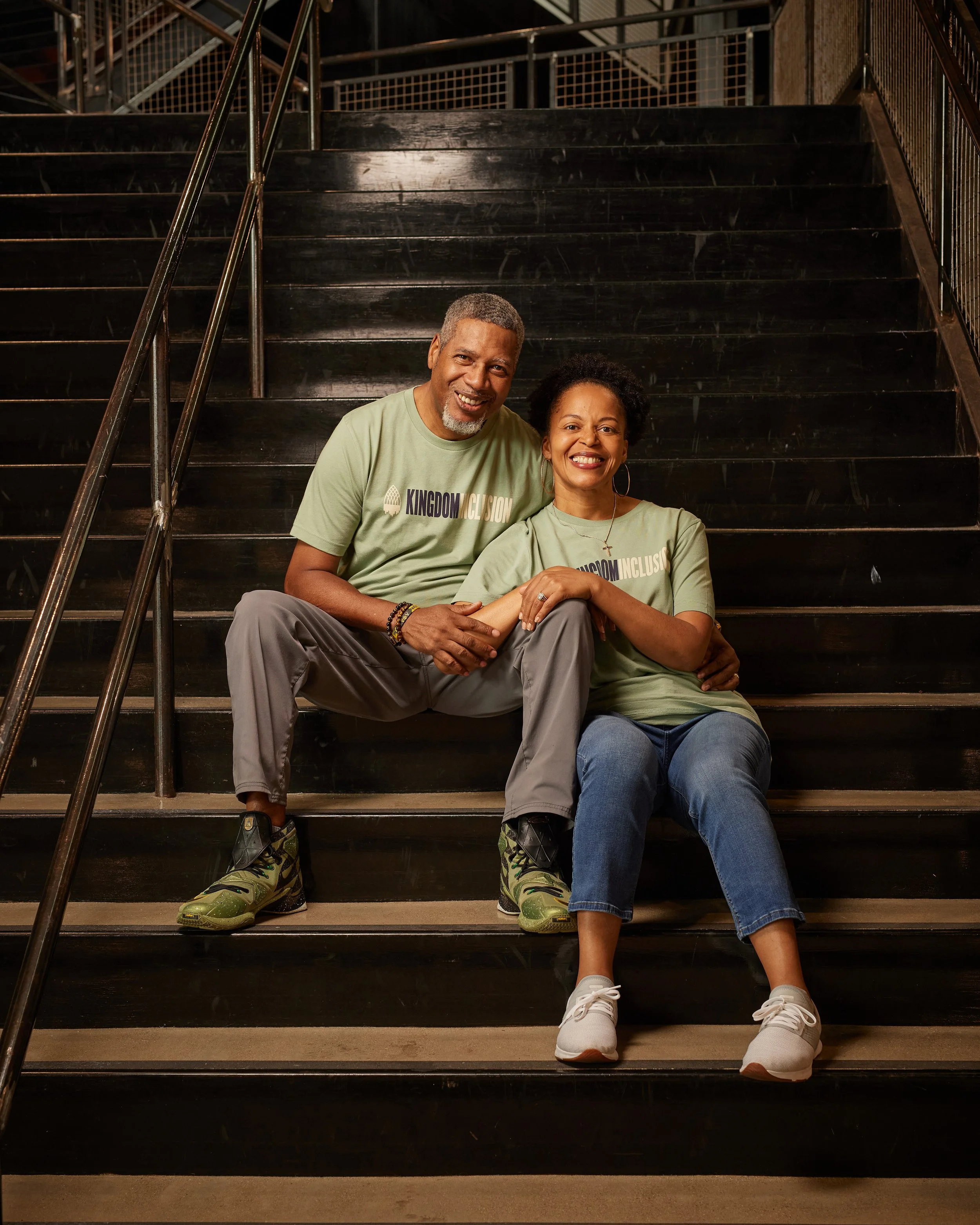 A smiling man and woman sitting on a staircase, holding hands, wearing matching green T-shirts with the words 'KINGDOM INCLUSION.'
