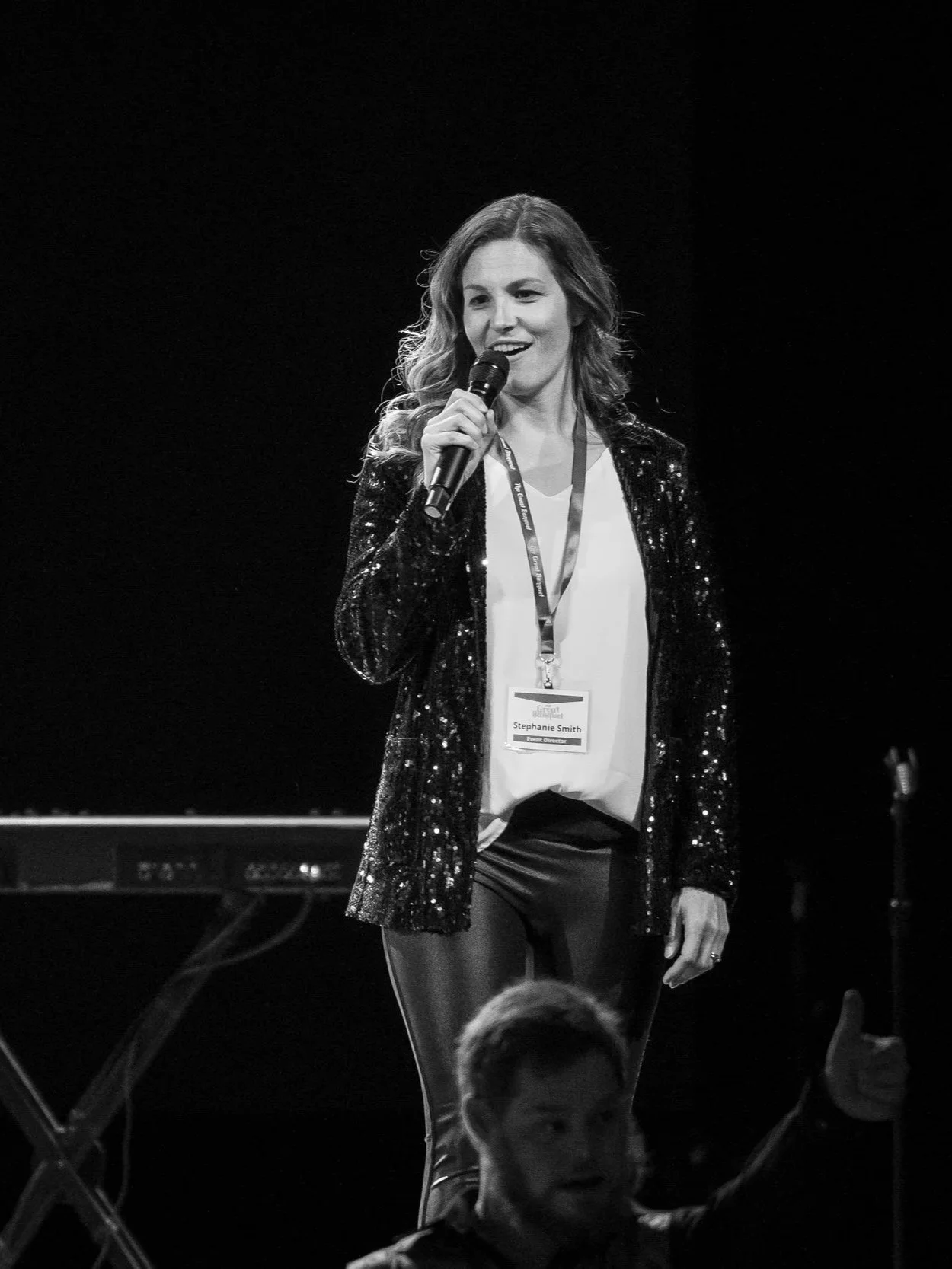 A woman speaking into a microphone on stage, wearing a black sequin blazer and a conference name tag that reads 'Stephanie Smith'.