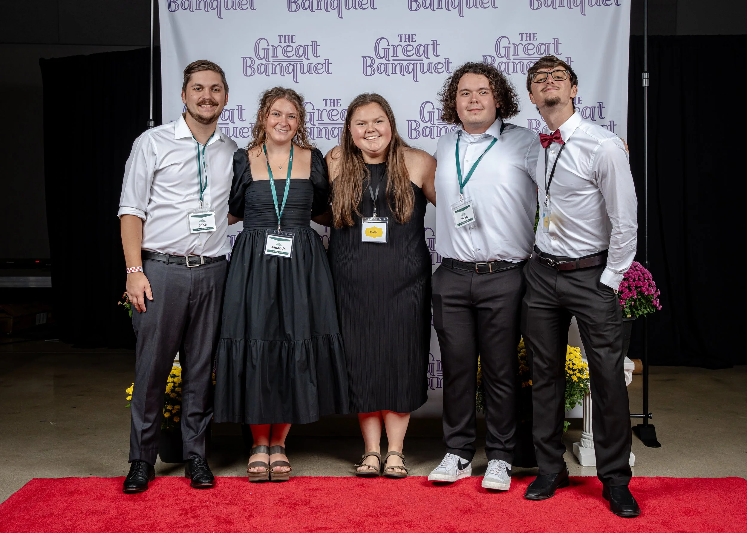 Five young adults standing on a red carpet in front of a backdrop that reads 'The Great Banquet.' They are dressed semi-formally, smiling, with name tags around their necks. The group includes three men and two women, with decorative flowers on either side of the group.