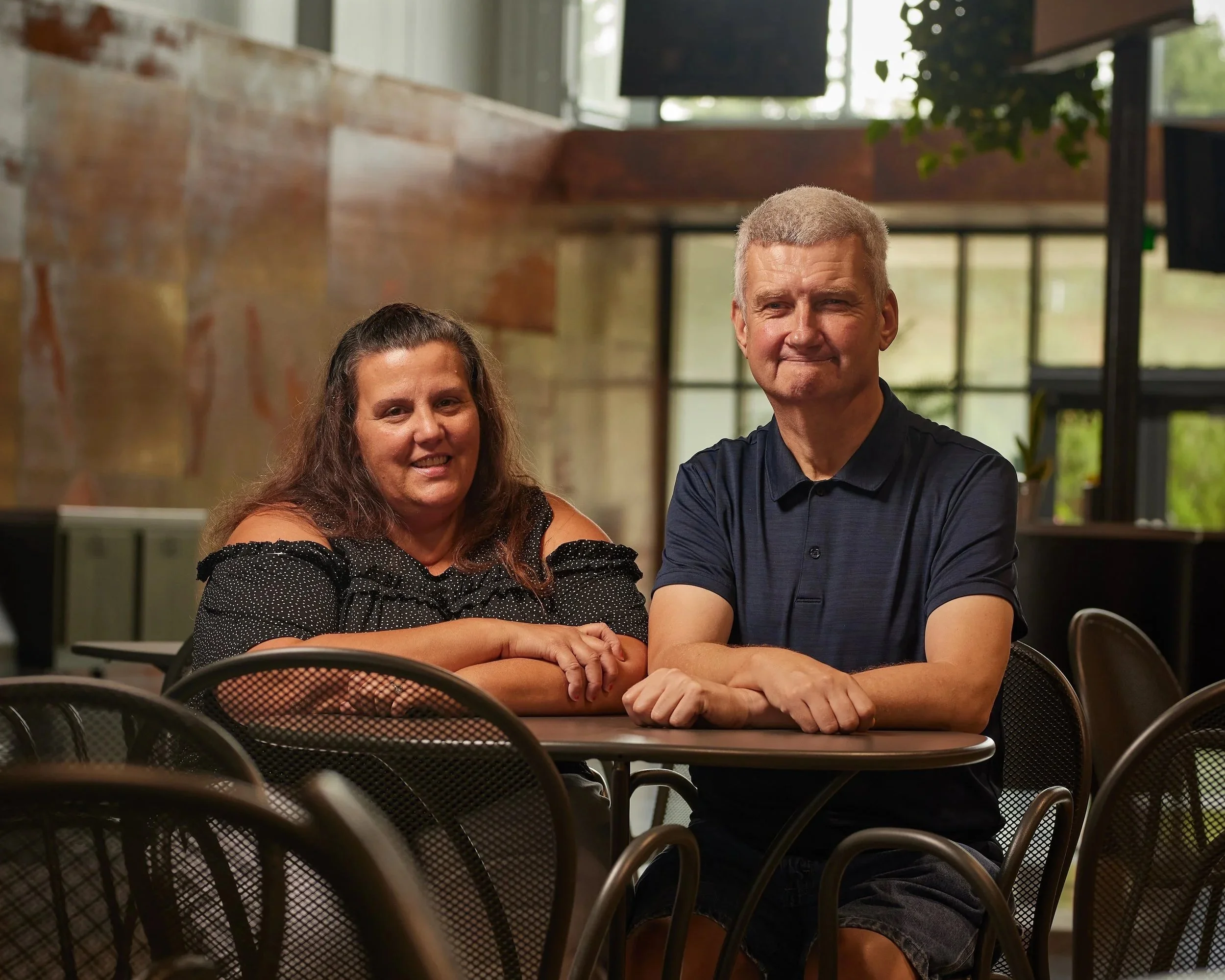A woman and a man sitting at a table indoors, smiling at the camera, with a modern rustic background.