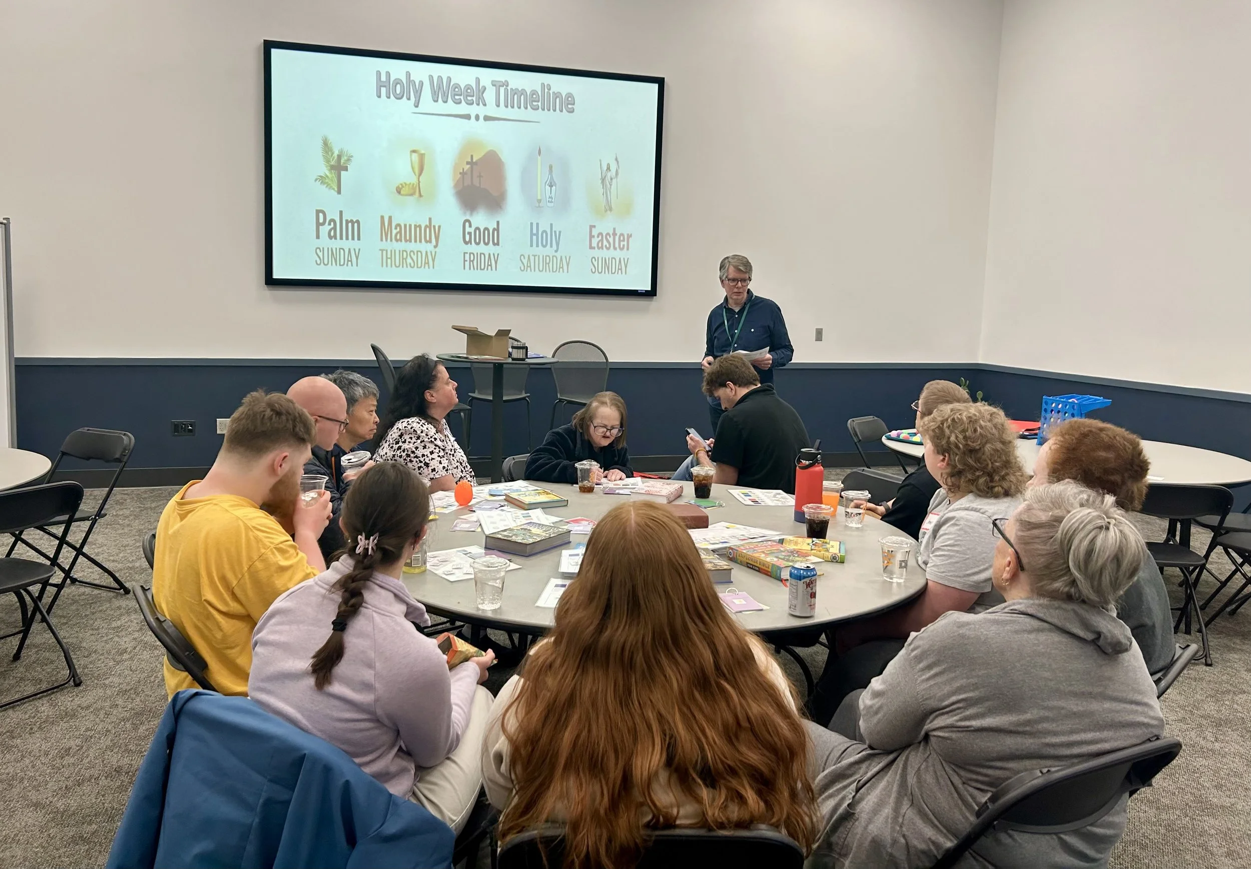 A group of people sitting around a large round table in a conference room, attending a presentation about the Holy Week timeline, with a man standing near a screen displaying the timeline with symbols and dates.