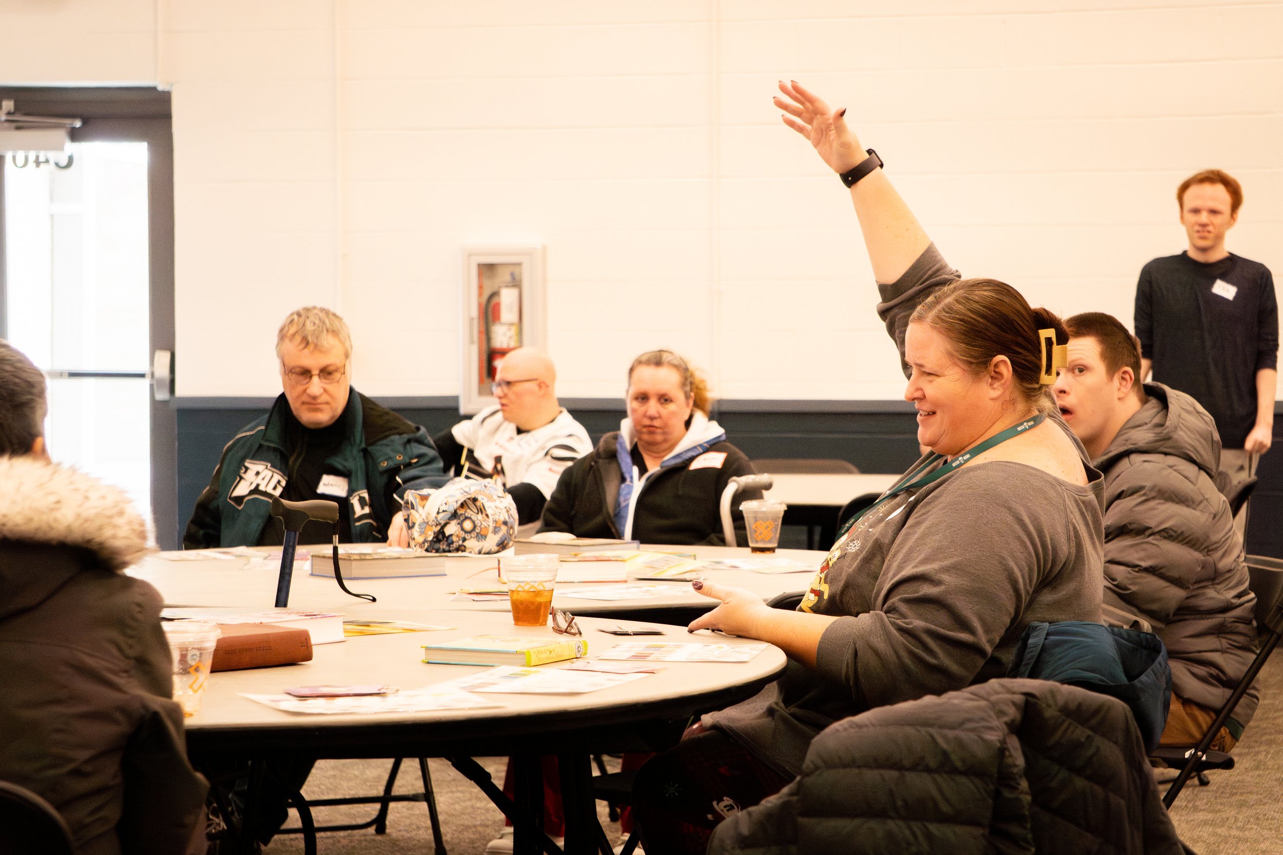 A group of people seated around a conference table, one woman raising her hand, in an indoor meeting room.