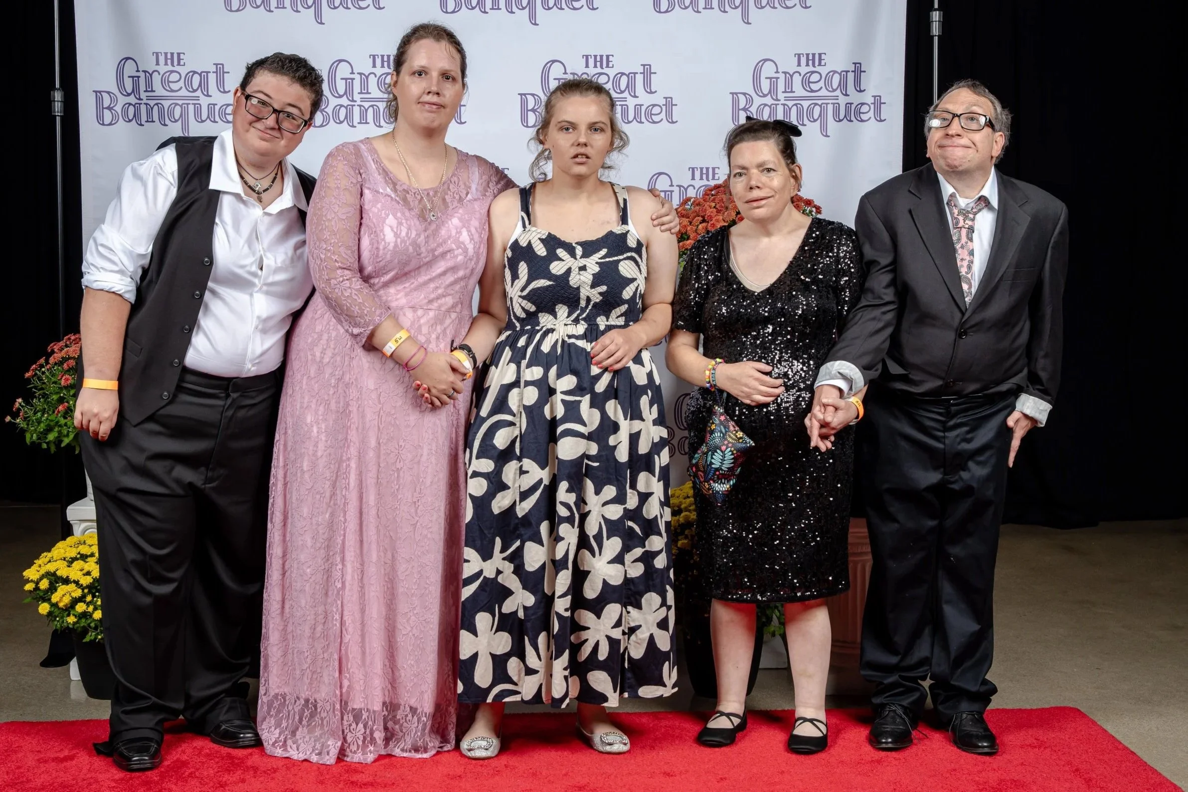 Five people standing on a red carpet at an event, with a backdrop that says 'The Great Banquet.' They are dressed in formal attire, and two of them are wearing wristbands.