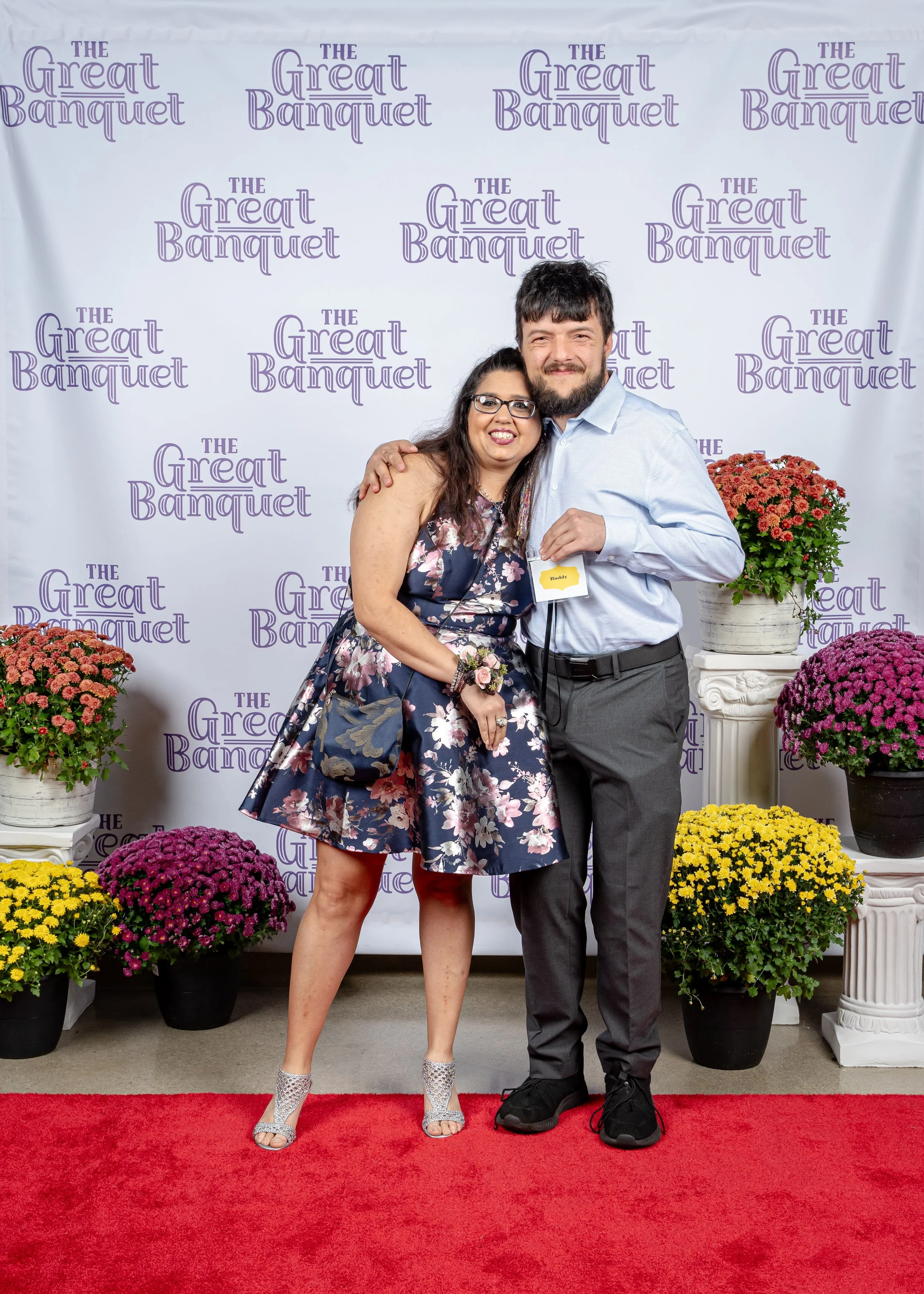 A woman in a floral dress and high heels standing next to a man in a light blue shirt and dark trousers, both smiling and hugging, standing on a red carpet in front of a backdrop with the words "The Great Banquet" and surrounded by colorful potted flowers.