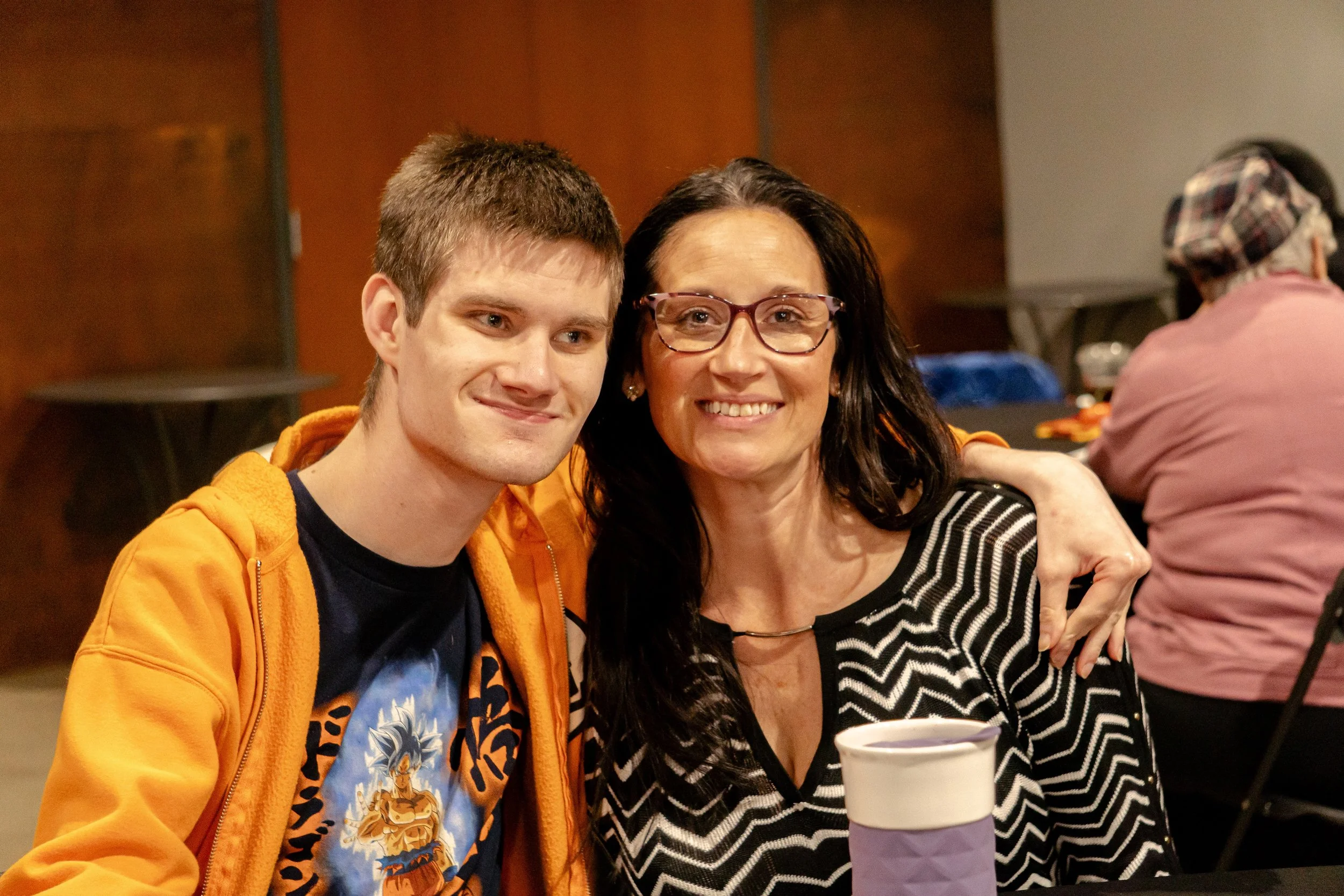 A young man and an older woman sitting together at a table, smiling, with a purple cup in front of them, in a room with people in the background.