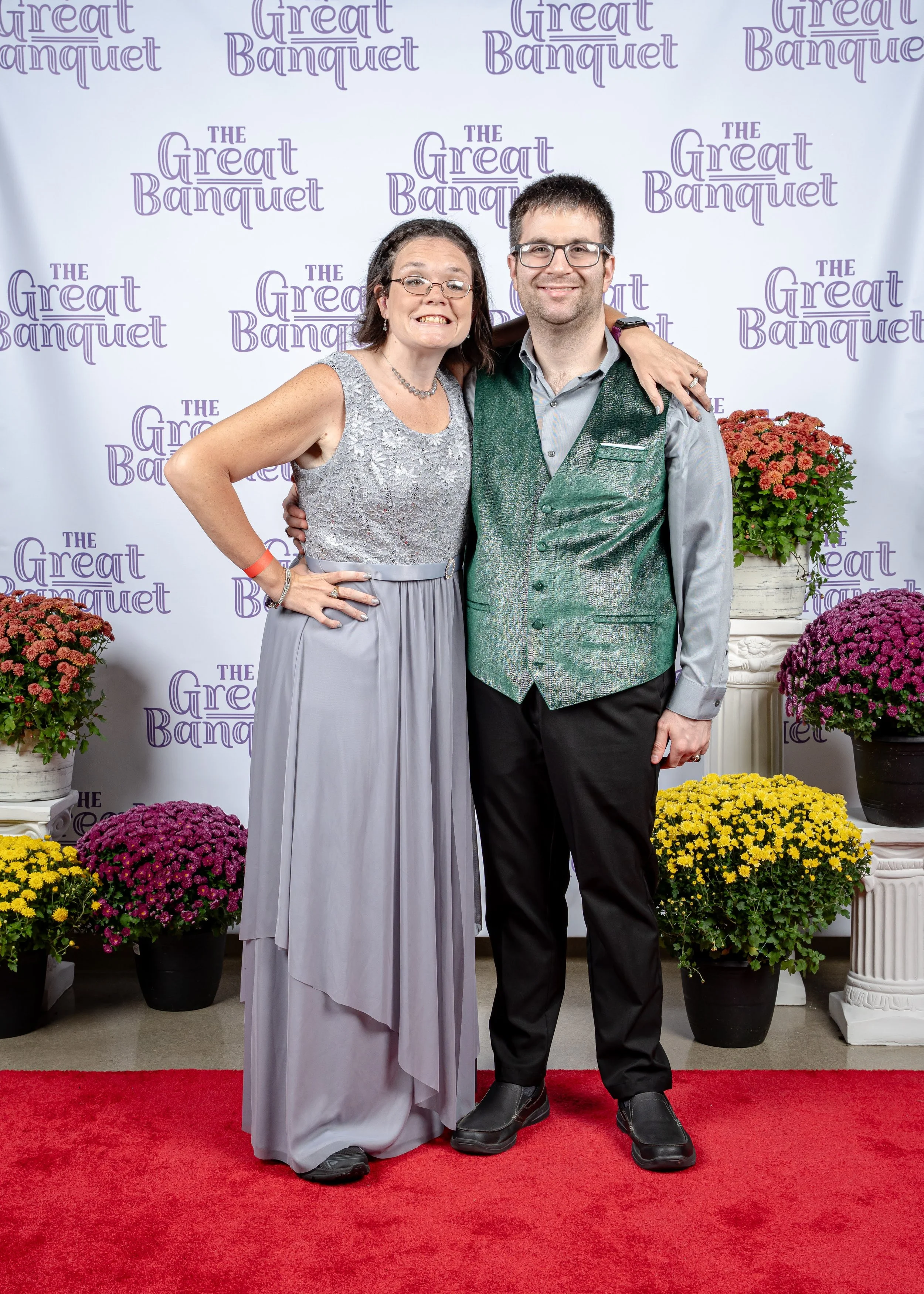 Two people, a woman and a man, standing on a red carpet with arms around each other, smiling at the camera. Behind them, a backdrop with the phrase 'The Great Banquet' in purple text and several flower arrangements in pots of pink, yellow, and purple.