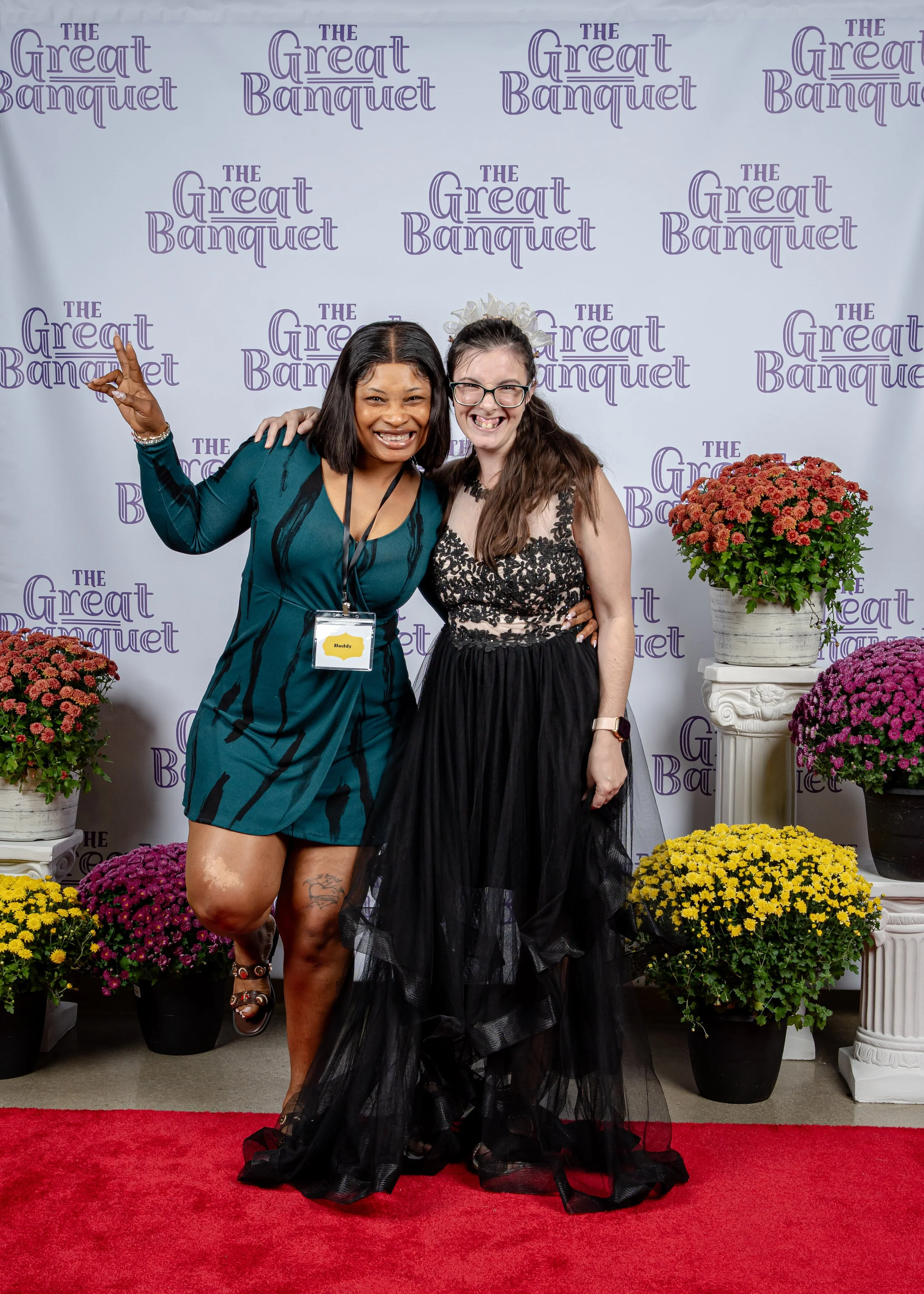 Two women posing on a red carpet in front of a backdrop with 'The Great Banquet' written in purple. One woman is wearing a teal dress with black patterns, has a name tag, and is making a peace sign. The other woman is dressed in a black lace and tulle gown, wearing glasses, and has a white floral headpiece, smiling and standing close together. Potted flowers surround them.