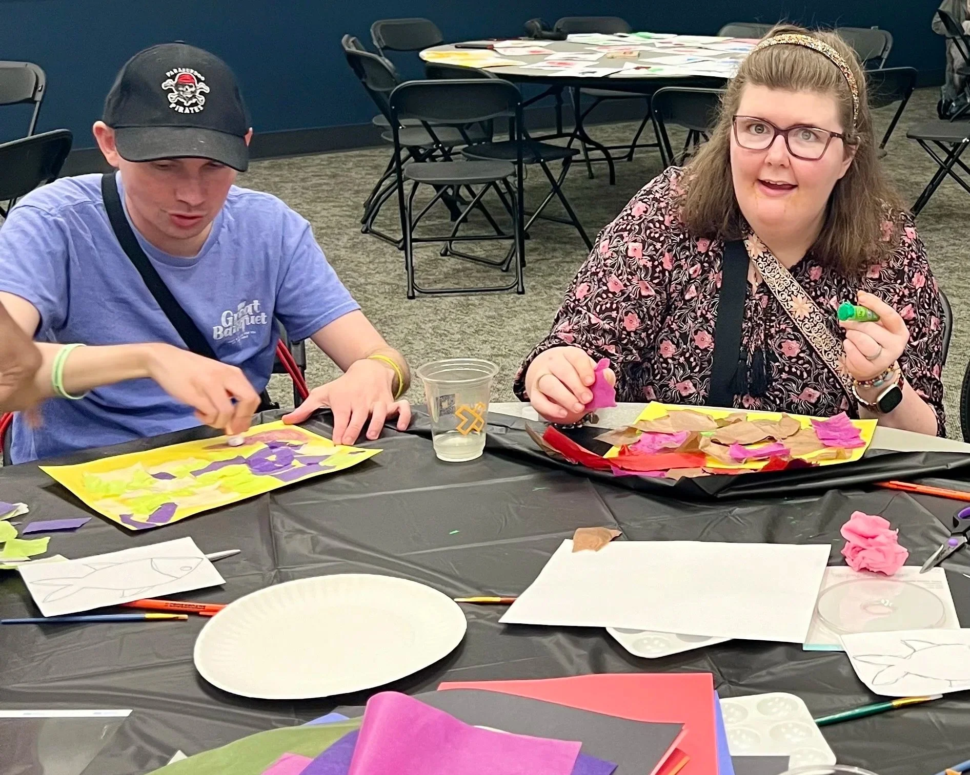 Two women sitting at a table engaged in arts and crafts activities, working with colorful tissue paper and glue, with art supplies scattered on the table.