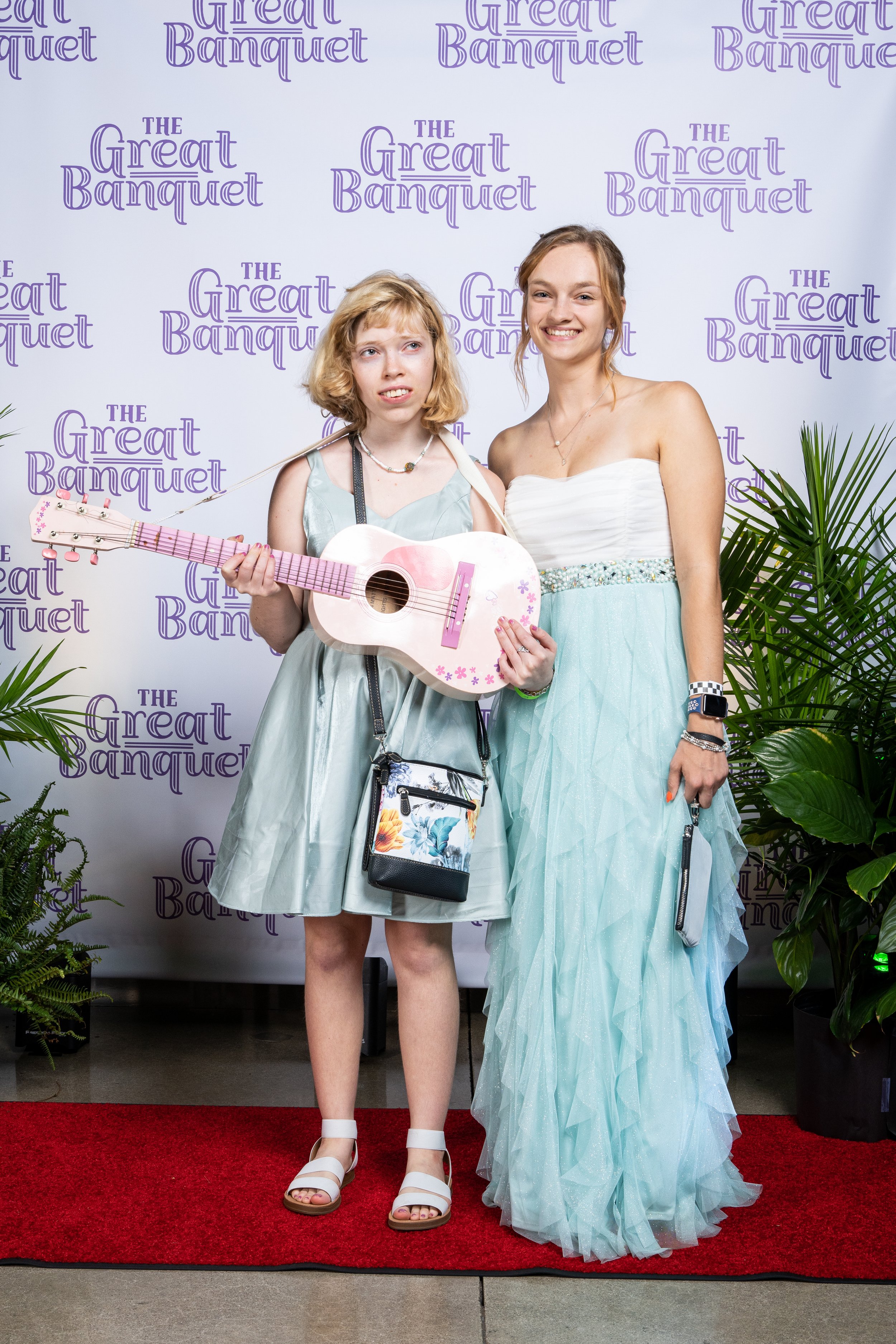 Two young women standing on a red carpet at an event called 'The Great Banquet,' with one of them holding a pink guitar. They are dressed in light-colored dresses, and there are green plants on either side of them.