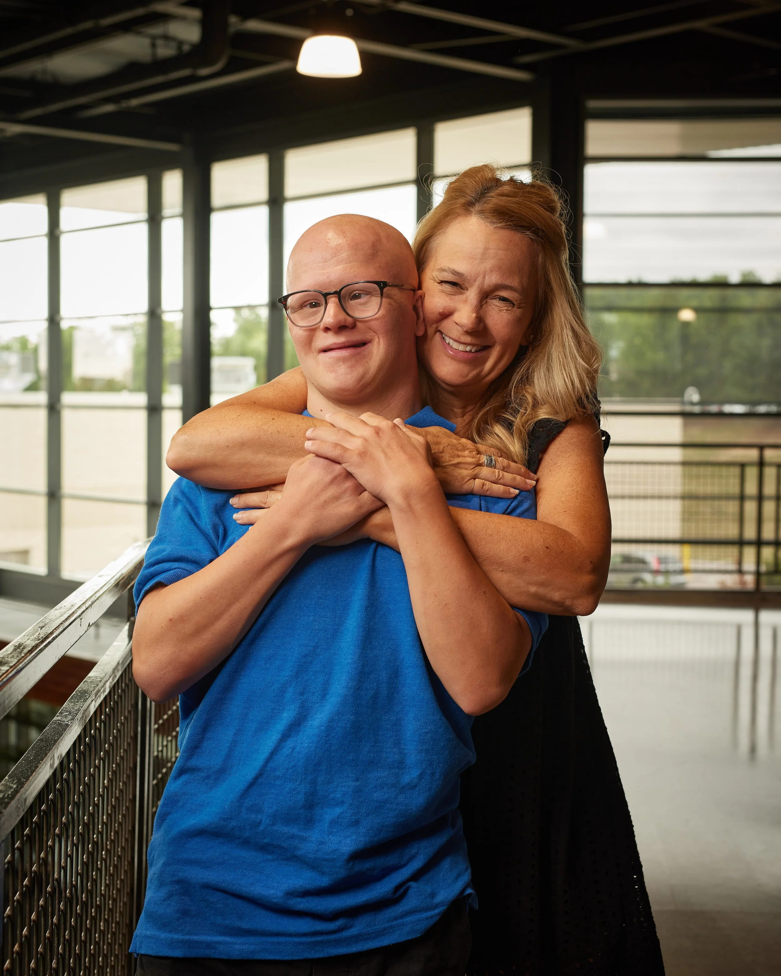 A woman hugging a young man with a shaved head and glasses from behind indoors, both smiling.