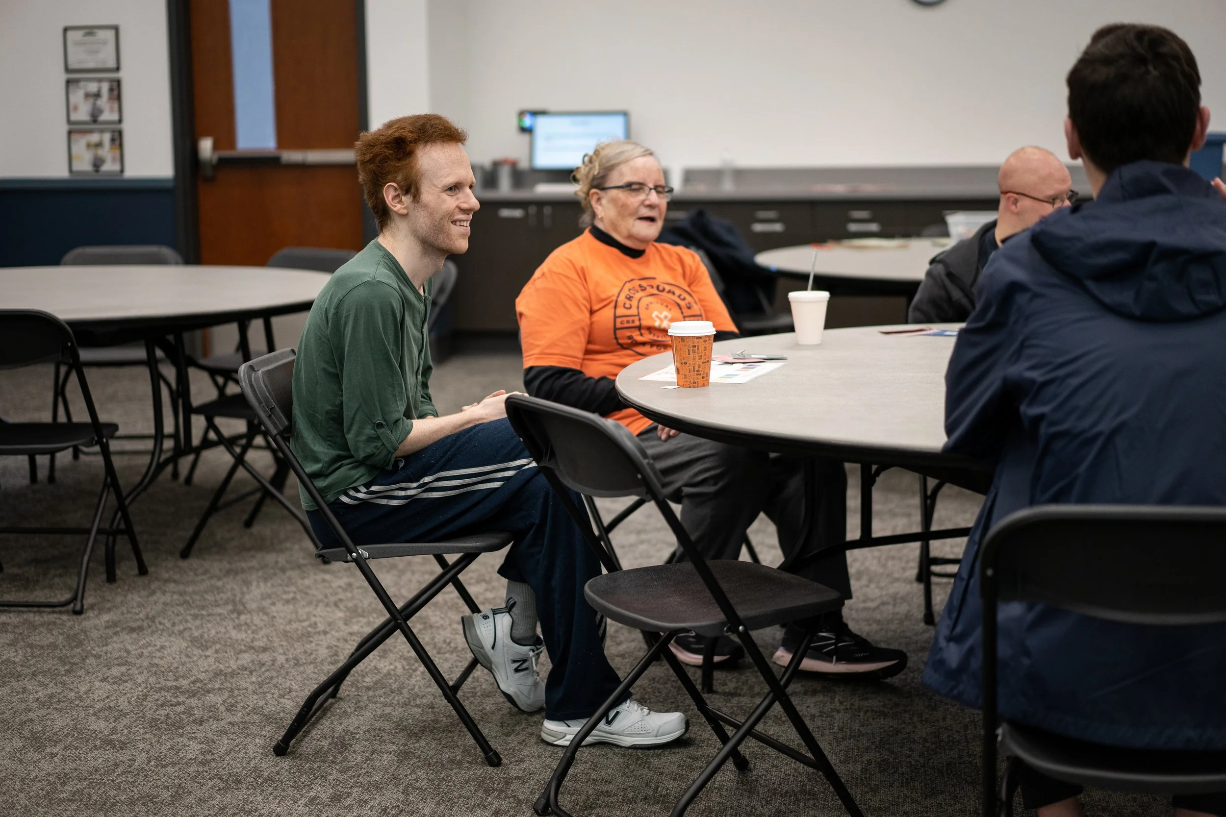 People sitting at round tables in a room, engaged in conversation. One woman is wearing an orange shirt, and a man in a green shirt is smiling. The room has a counter with a computer in the background.