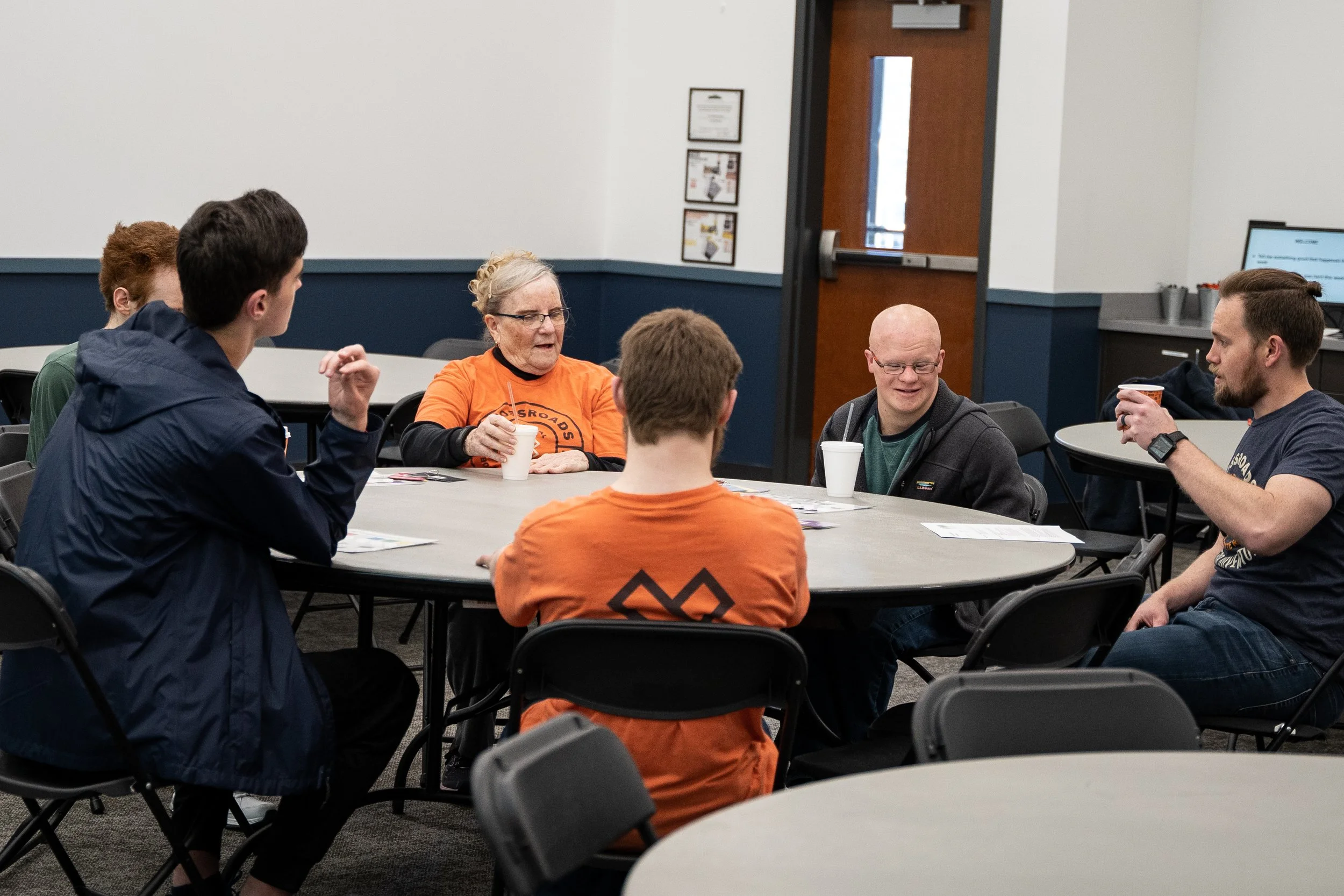 Group of six people sitting around a round table in a conference room, some wearing orange shirts, chatting and drinking beverages.