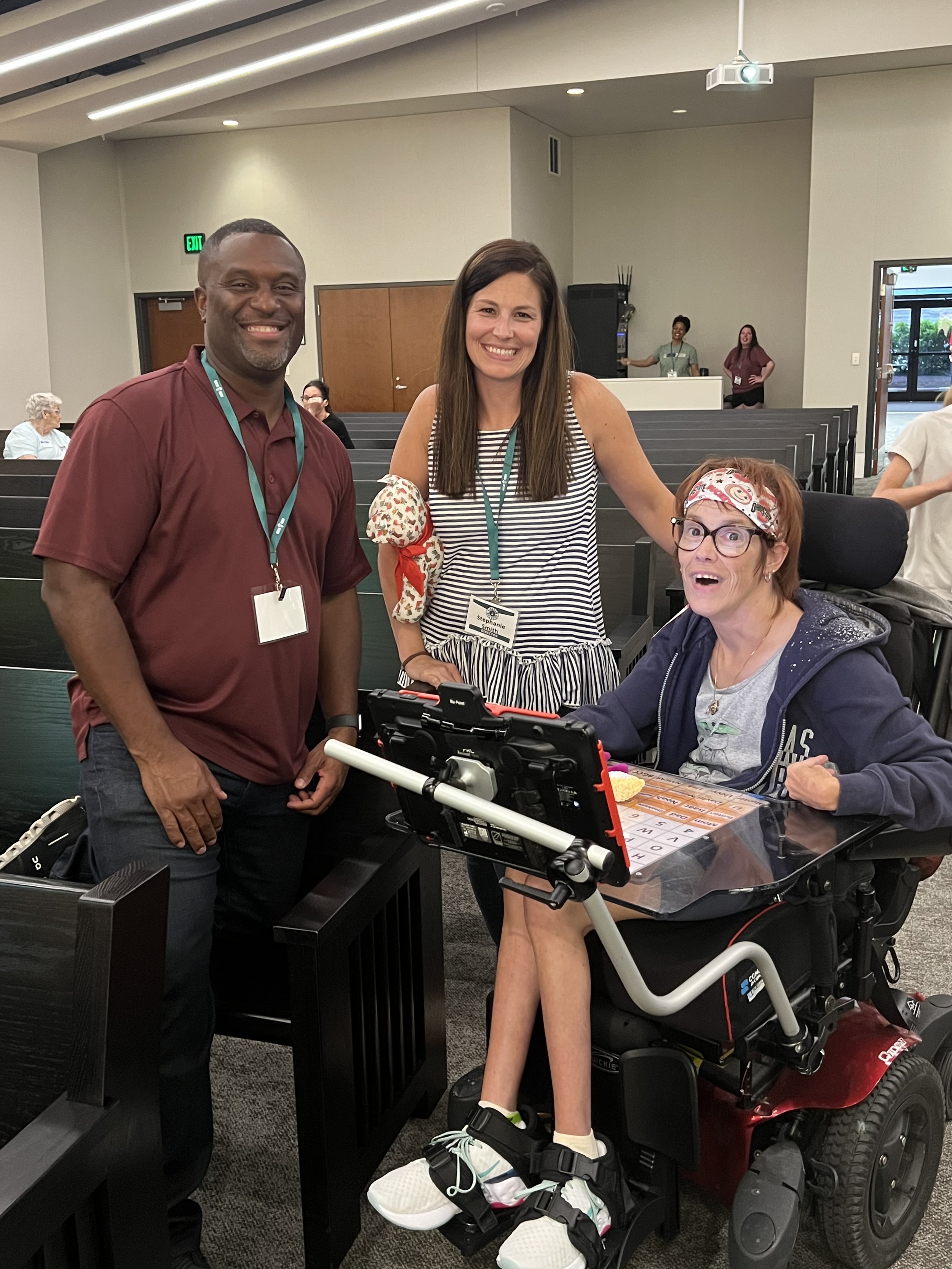 Three people smiling at the camera in a room with chairs, one woman in a motorized wheelchair and two standing women, one in a burgundy shirt and the other in a striped dress, with name tags around their necks.
