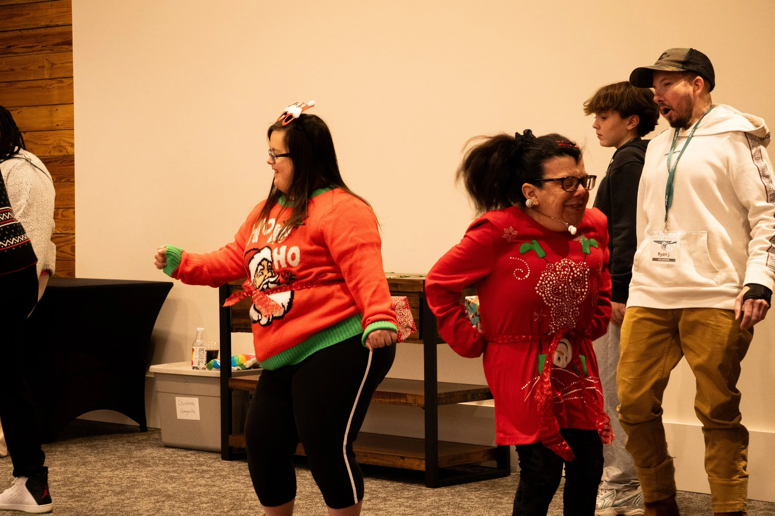 People celebrating at a Christmas event. Two women in Christmas sweaters are dancing, with one wearing a red sweater with a reindeer and the other wearing a red sweater with Christmas decorations. A man and a young boy are talking nearby.