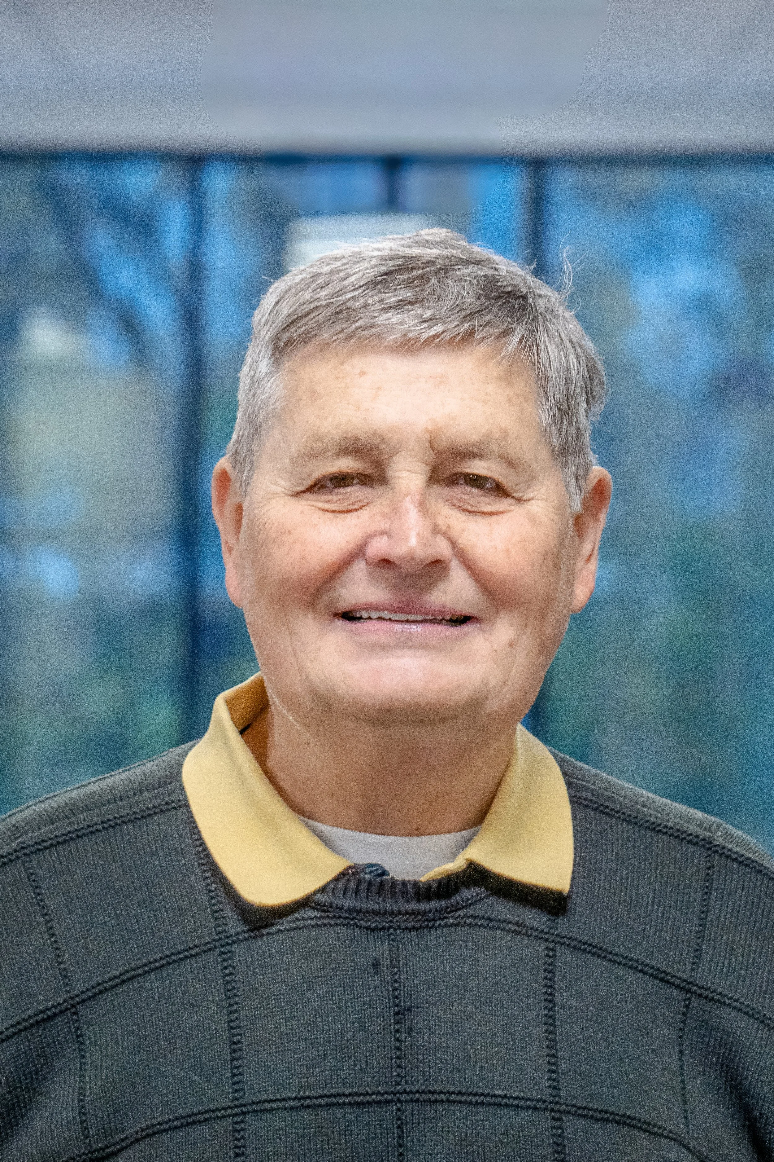 A smiling middle-aged man with short gray hair, wearing a dark sweater with a yellow collar, standing indoors.