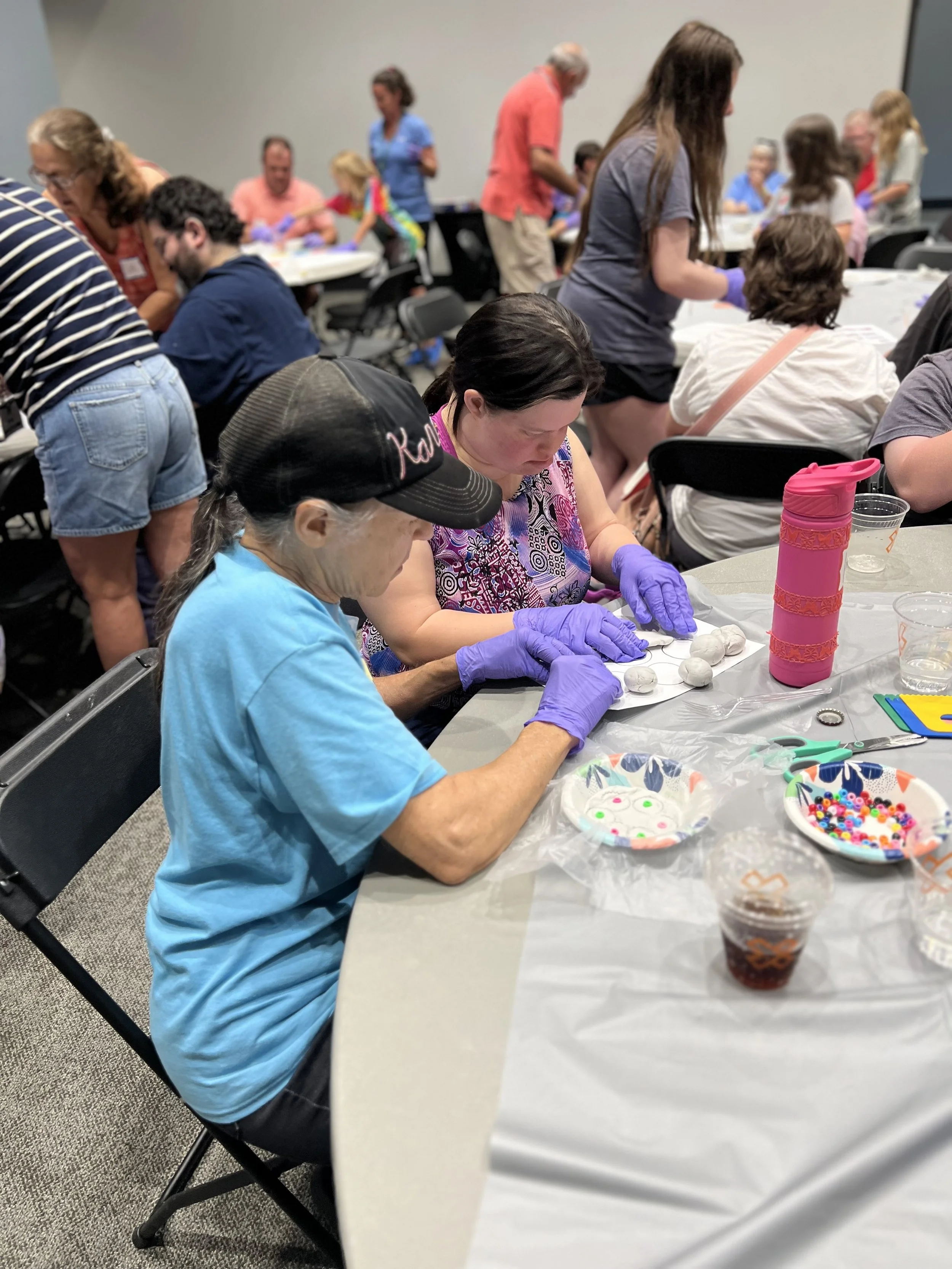 People at a craft workshop decorating spheres with buttons and markers, seated at tables with supplies, in a large meeting room.