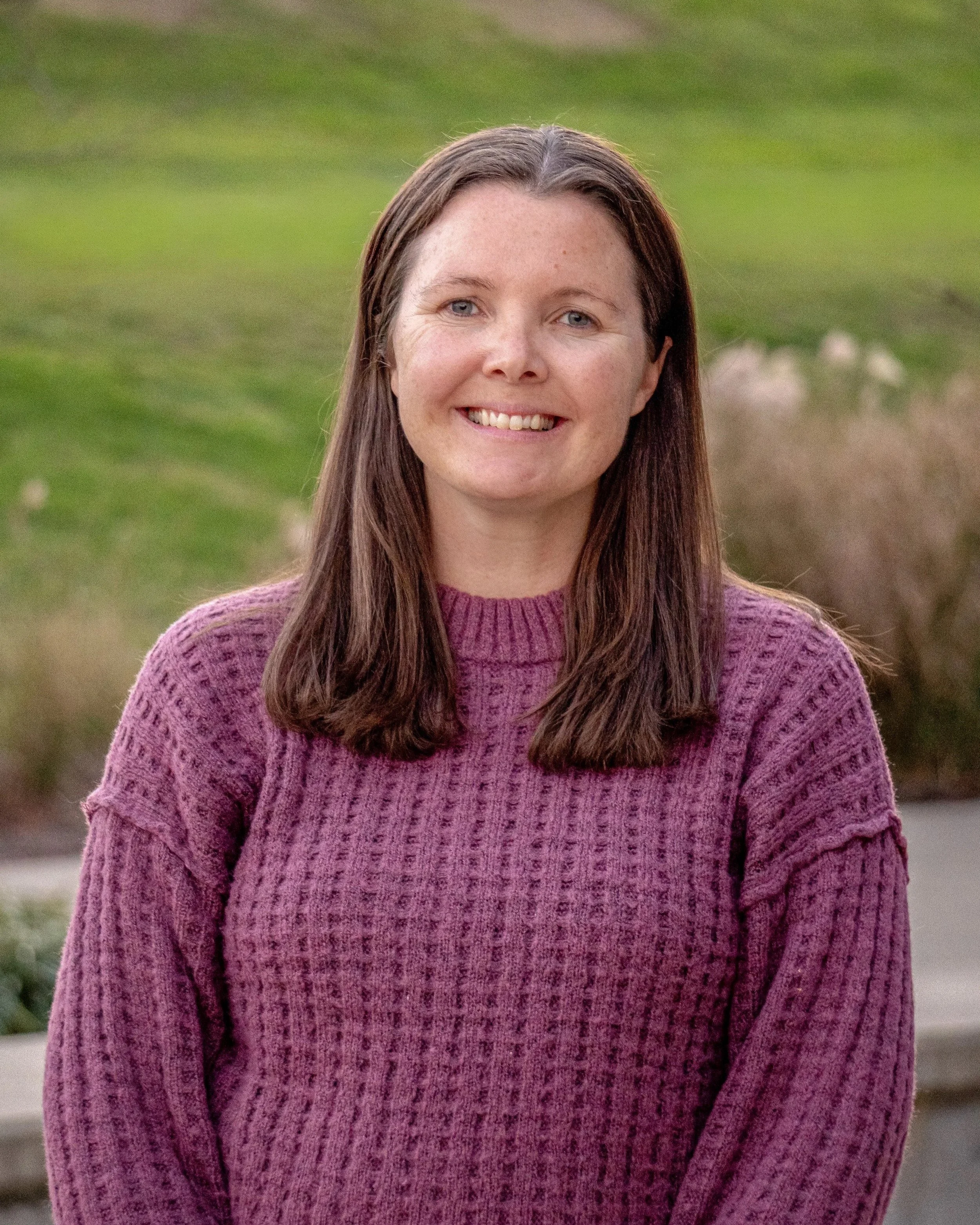 A woman with long brown hair wearing a pink sweater, smiling outdoors with a grassy area in the background.