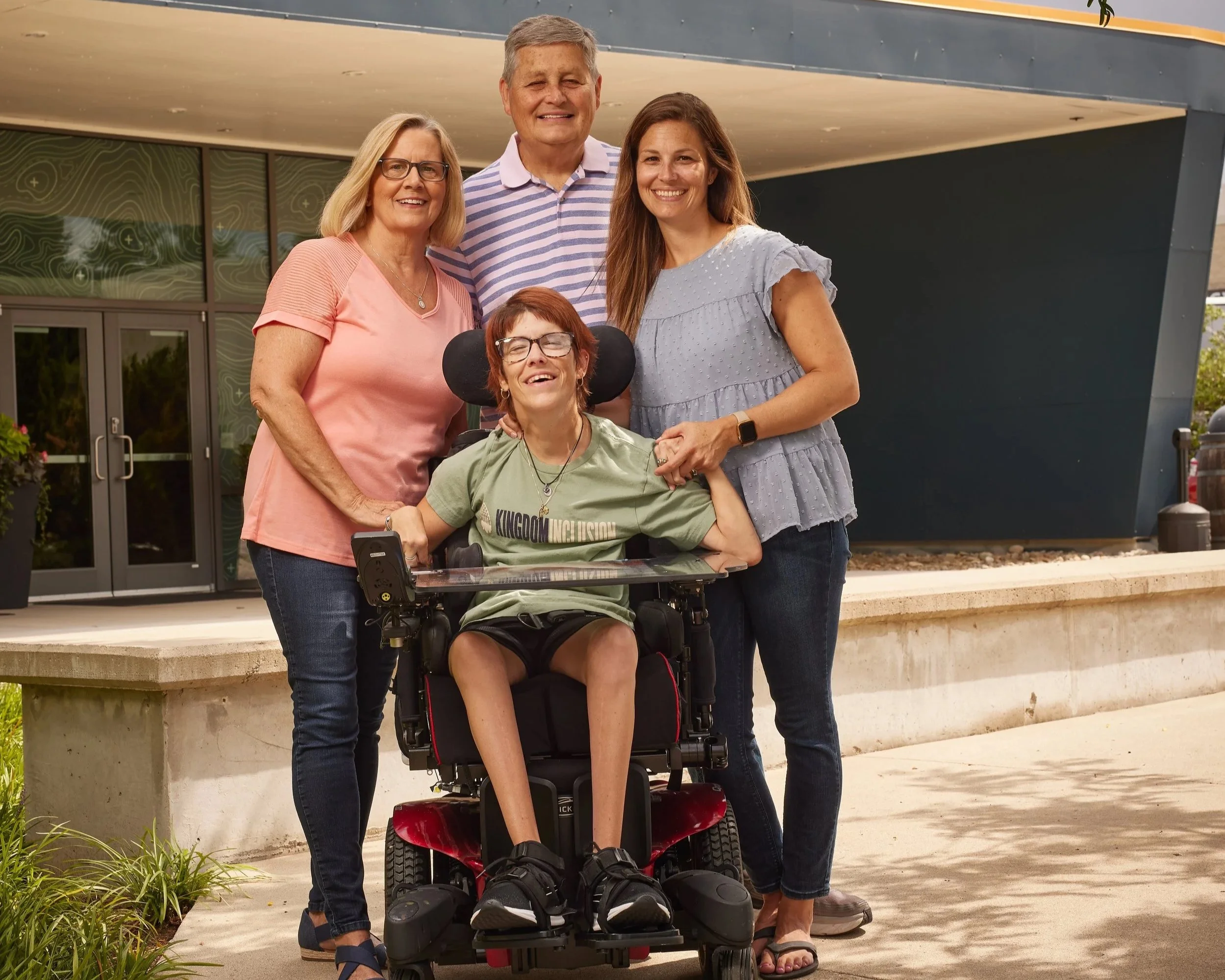A group of five people, including a young person in a motorized wheelchair and two women, standing outdoors in front of a modern building, smiling for a photo.