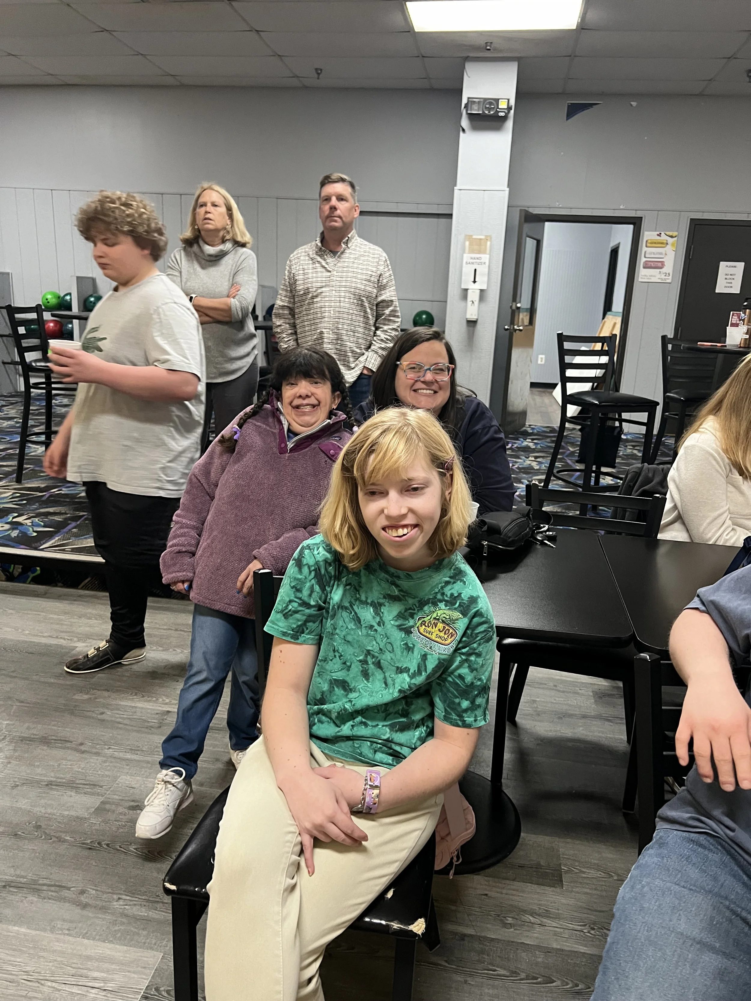 Group of five people in a restaurant or bowling alley, with three women and two children, smiling and posing for the camera. The setting has black chairs, tables, and a wall with a door in the background.