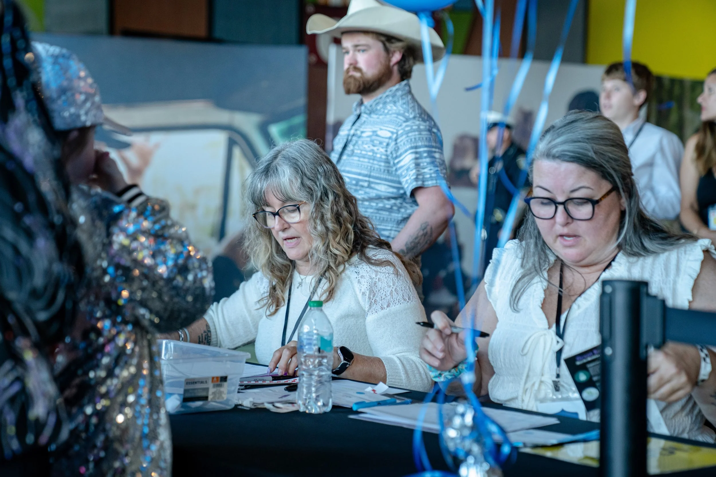 Two women with glasses and gray hair sitting at a table, writing or reading, with an elderly woman in the foreground, a man with a cowboy hat and beard in the background, and some people standing behind. The scene appears to be at an indoor event with blue and yellow decorations and blue streamers hanging down.