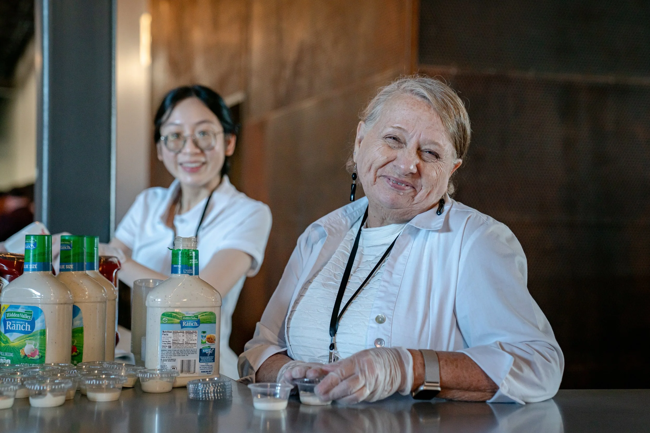Two smiling women, one with glasses, sitting at a table with bottles of ranch dressing and small containers of dipping sauce.