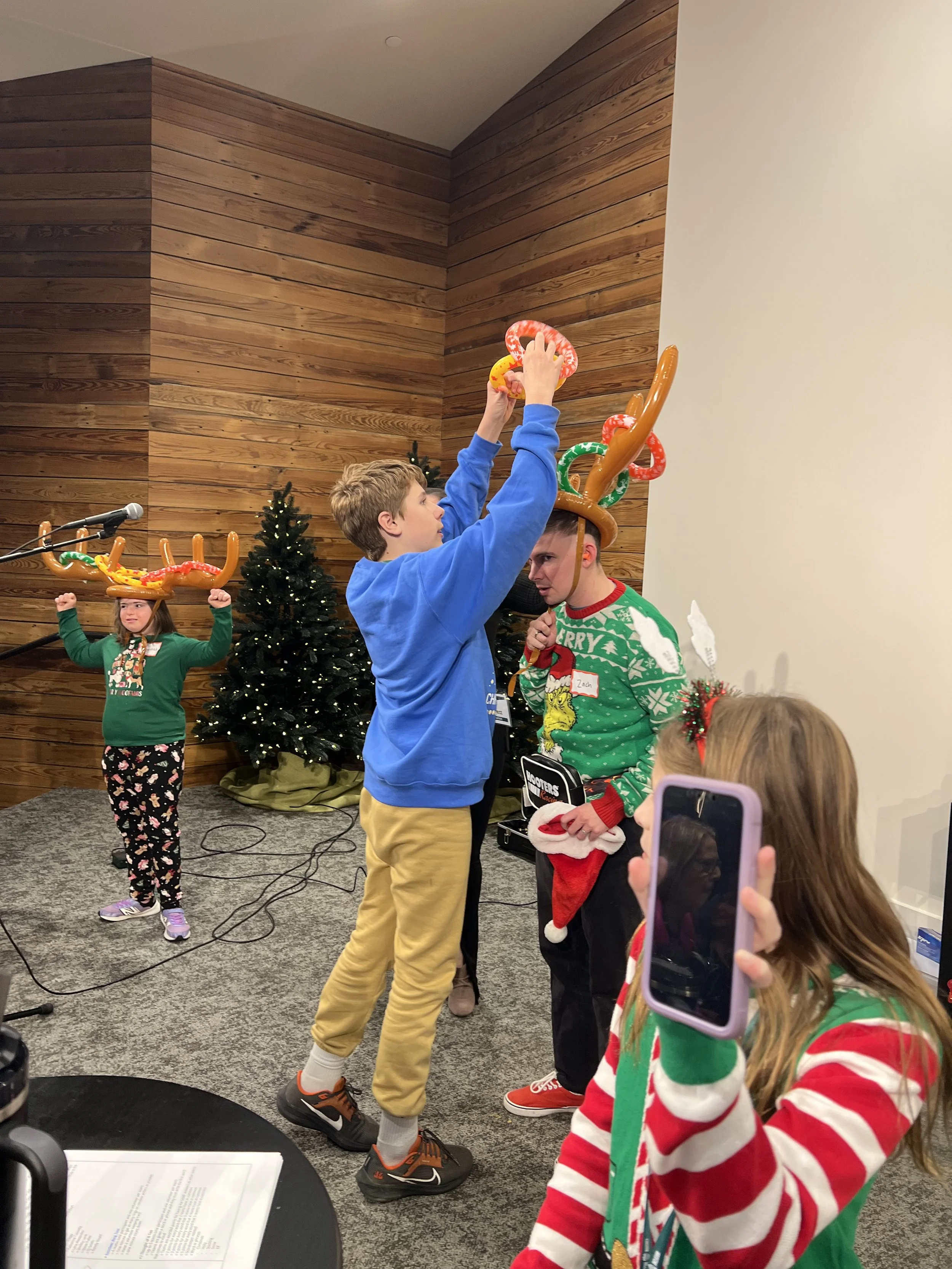 Children on a stage decorating Christmas hats with reindeer antlers during a holiday celebration, with Christmas trees in the background.
