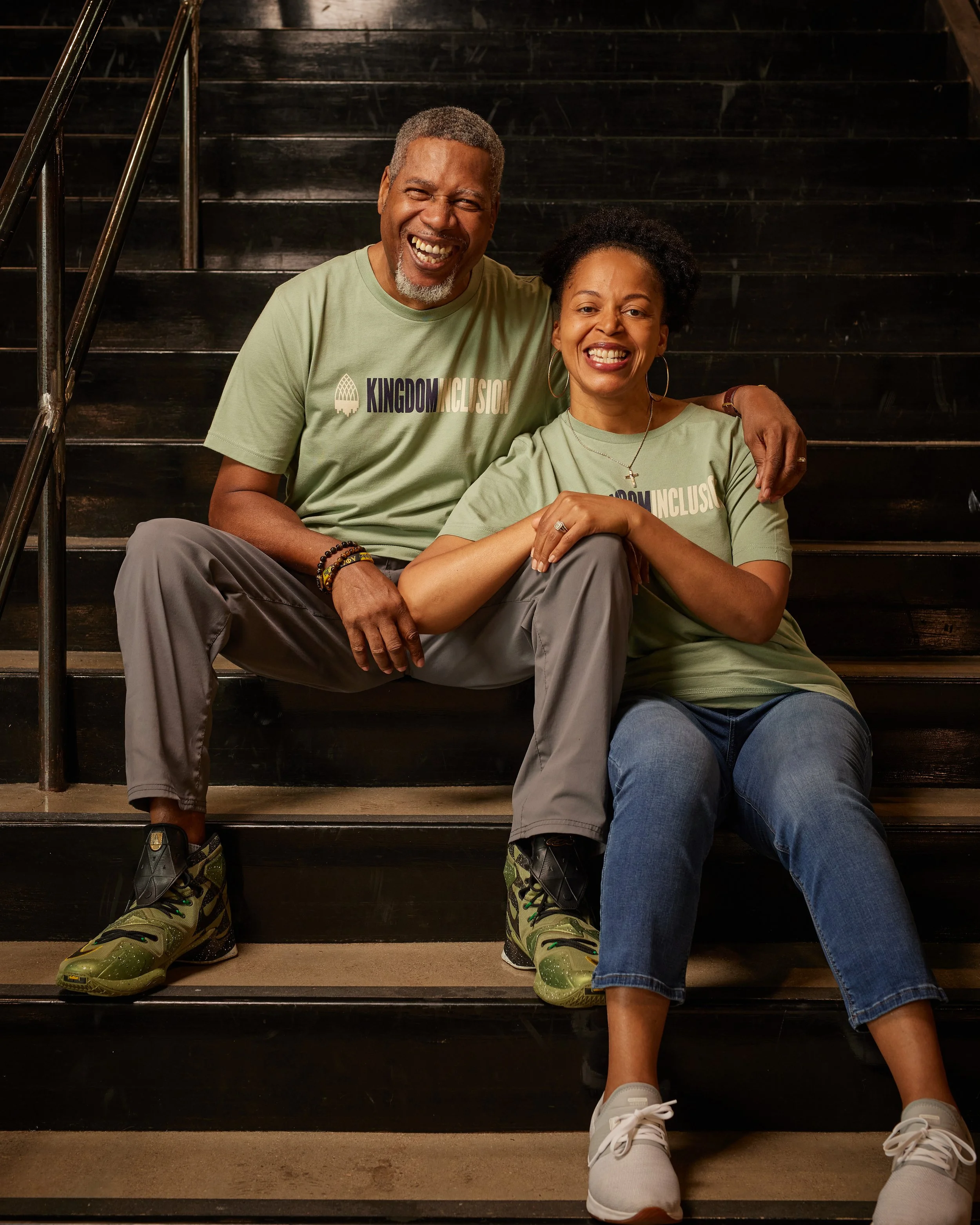A happy middle-aged African American man and woman sitting on dark wooden stairs, smiling and enjoying each other's company, both wearing matching green t-shirts with the words 'Kingdom Inclusion'.