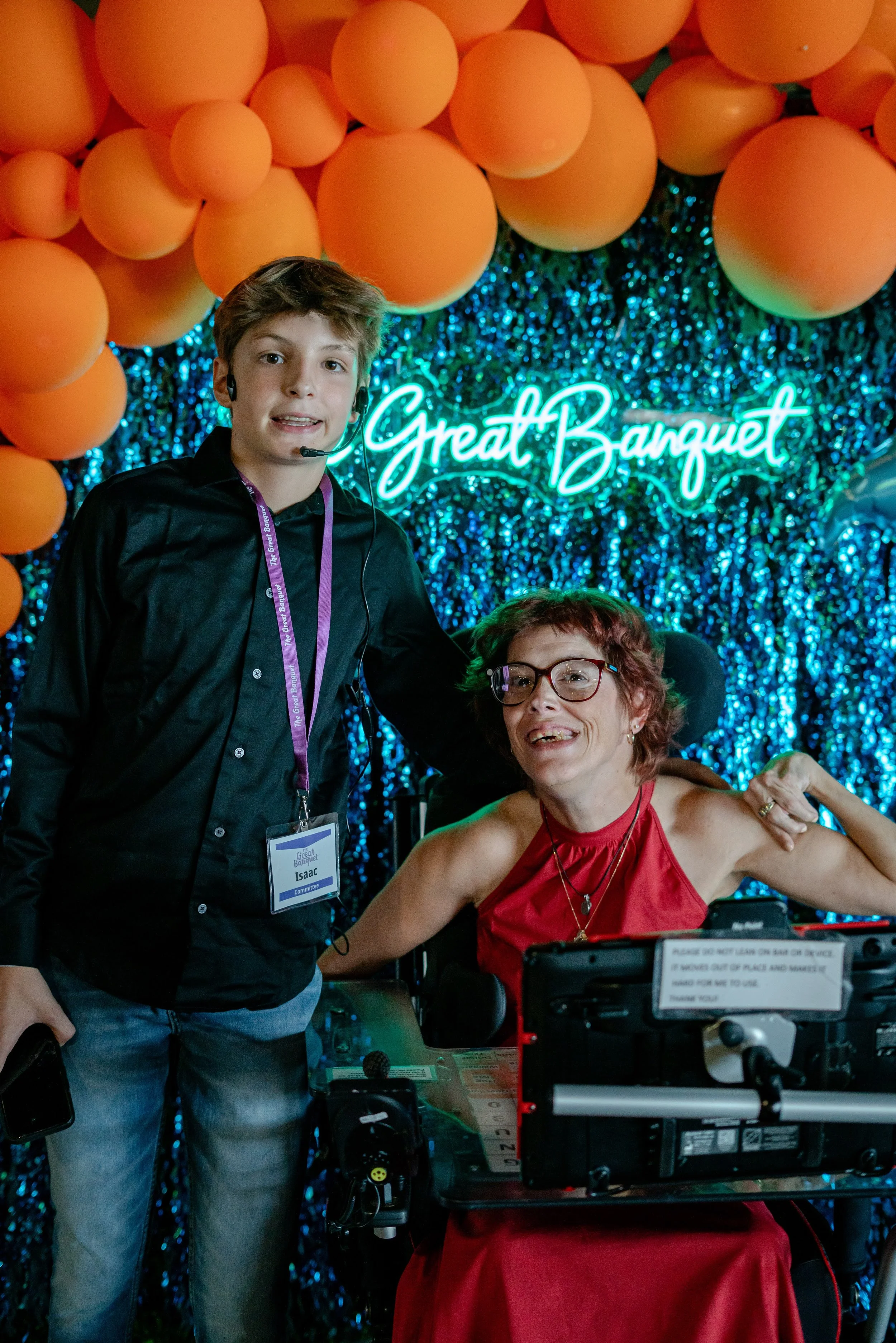 A young boy with a headset and a woman in a wheelchair, both smiling, at a festive event with a sign that reads 'Great Banquet' in neon lights behind them.
