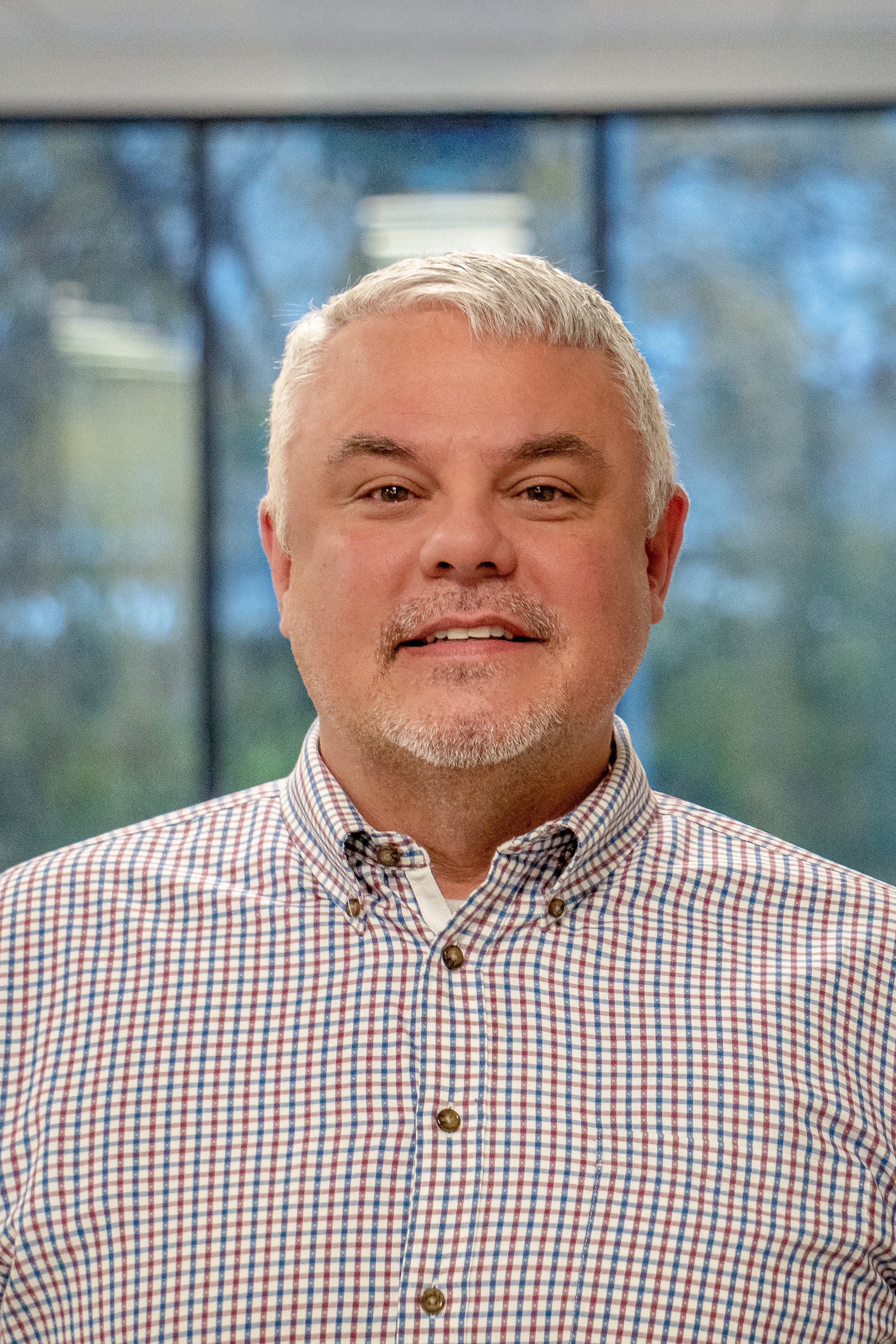 A middle-aged man with short gray hair, a goatee, and a mustache, wearing a checkered button-down shirt, standing indoors in front of a window with a blurred outdoor background.