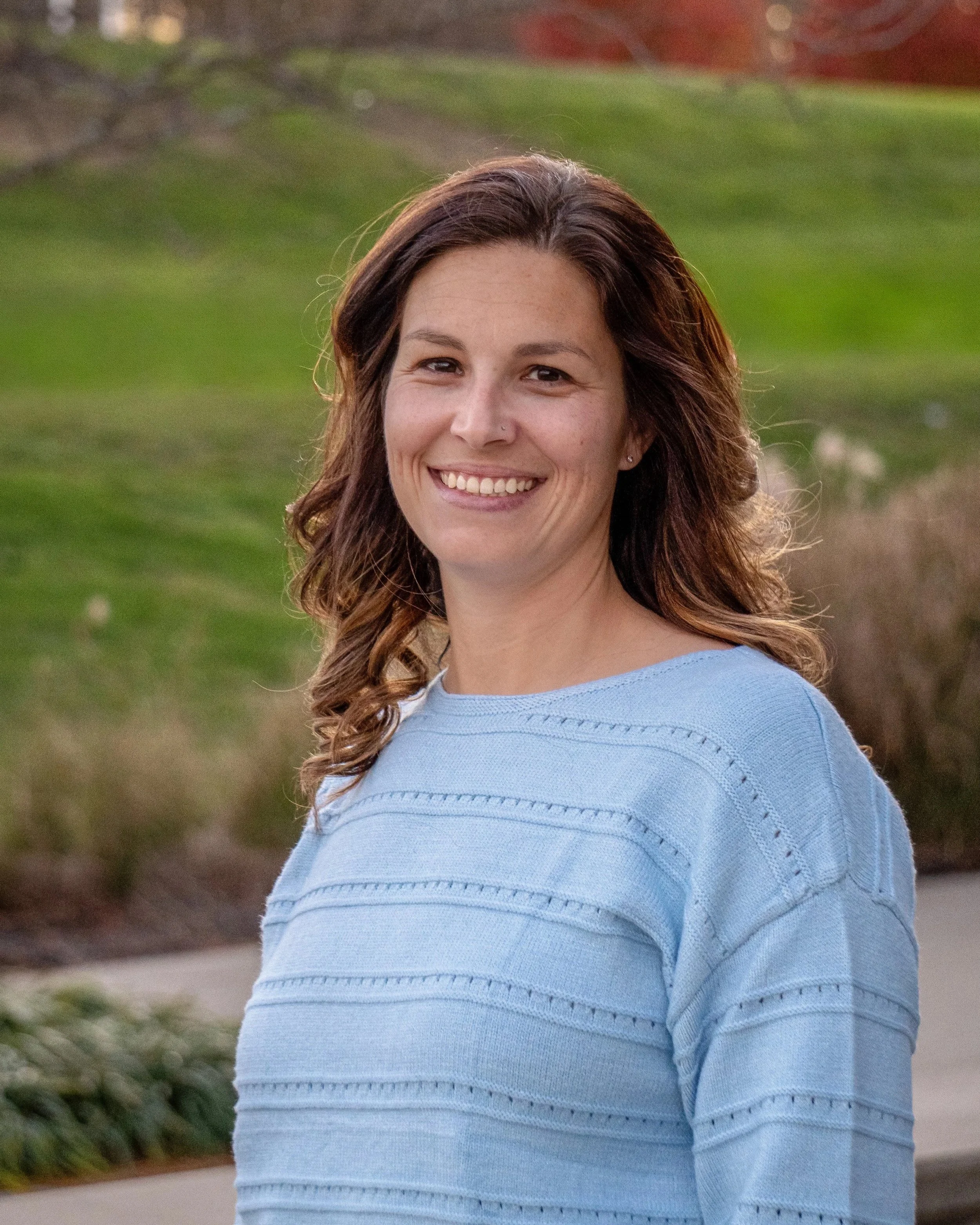 A woman with wavy brown hair smiling outdoors, wearing a light blue sweater, with a background of grass and trees.
