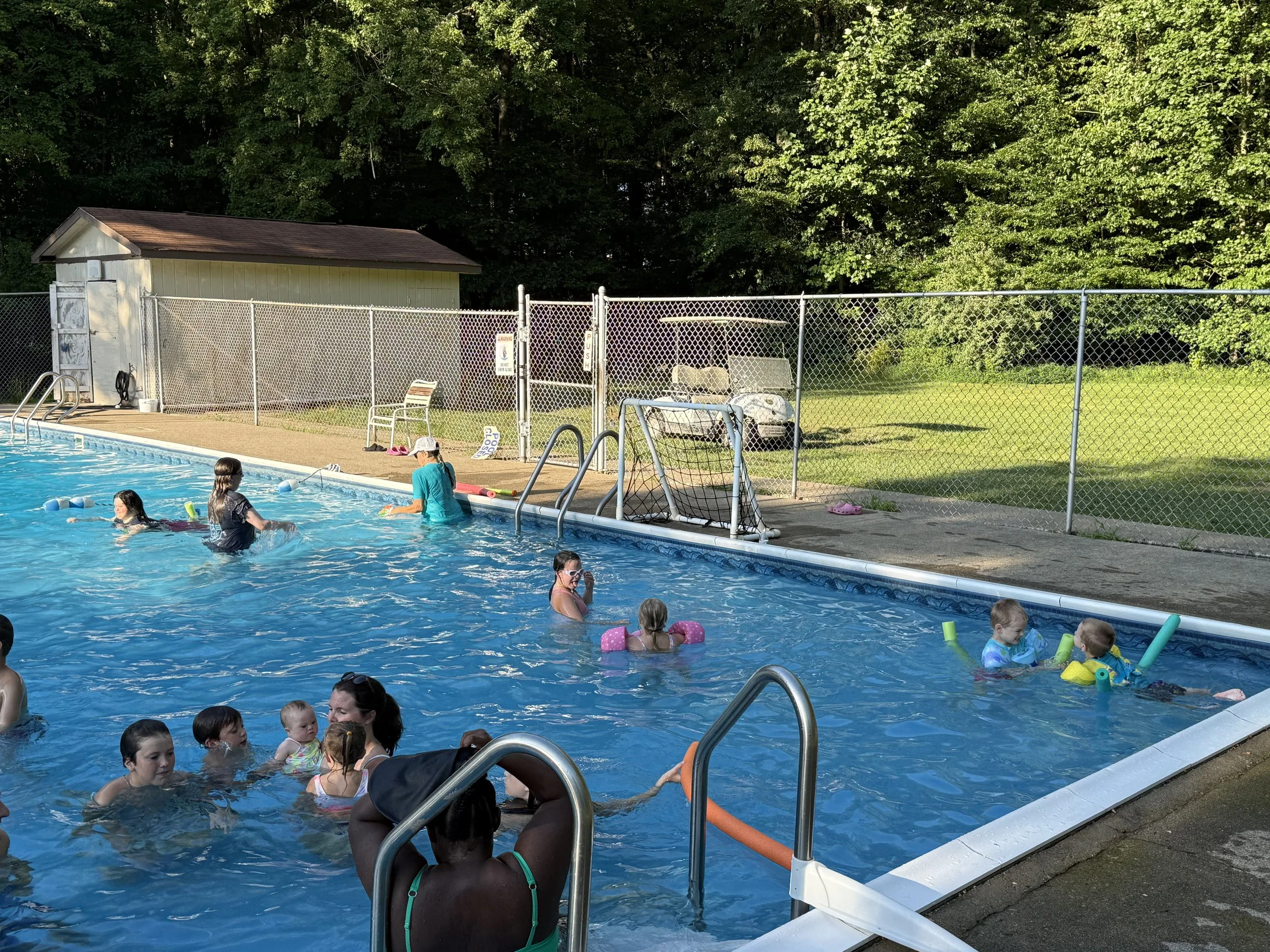 People swimming and playing in an outdoor community pool during daytime, surrounded by a chain-link fence and lush green trees.