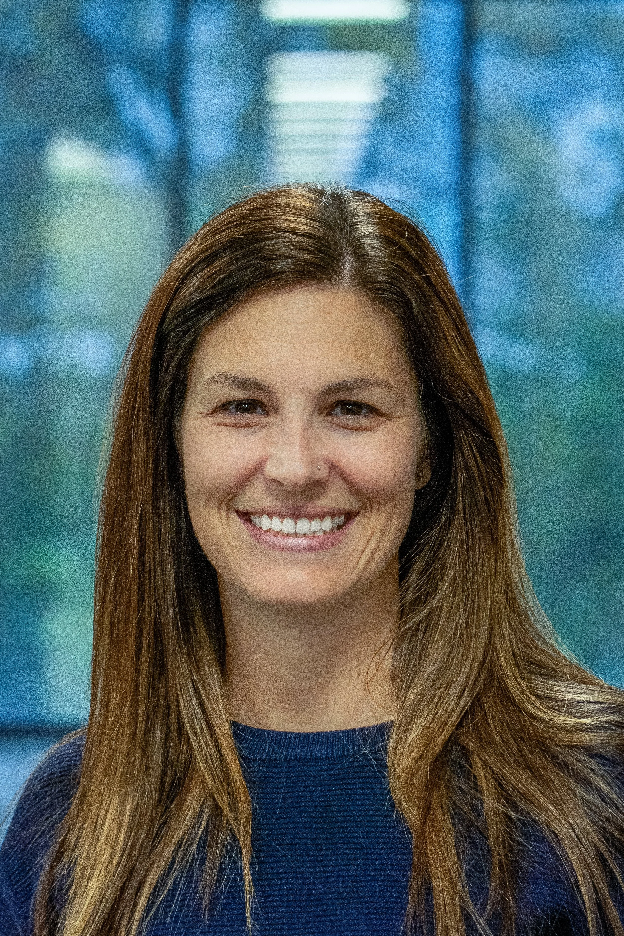 Smiling woman with long brown hair, wearing a dark blue top, posing in front of a blurred blue indoor background with windows.