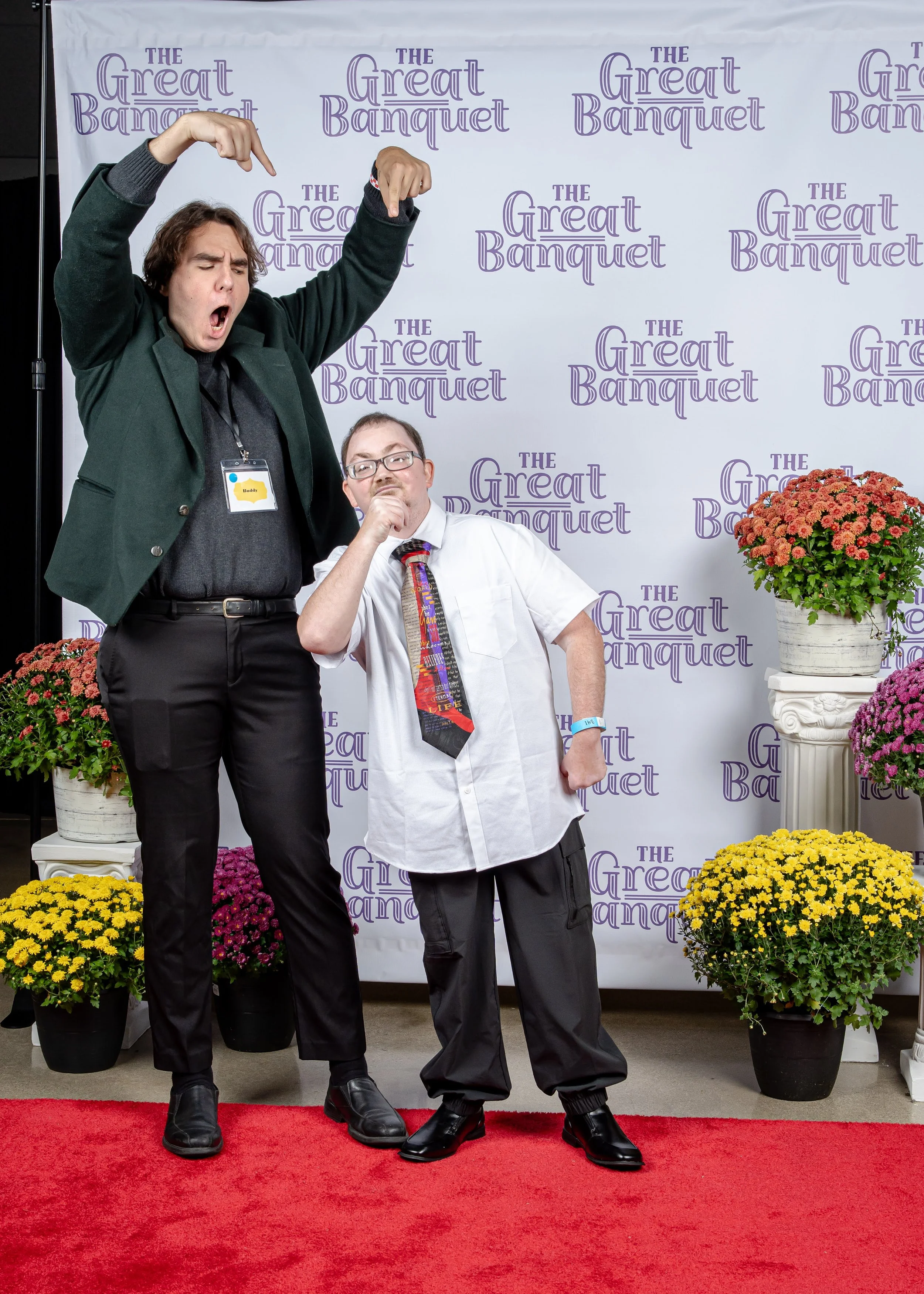Two men posing at an event with a backdrop that says 'The Great Banquet'. One is tall with long hair, wearing a dark green blazer and black pants, making a dramatic gesture. The other is shorter, wearing glasses, a white shirt, a colorful tie, and black shoes, striking a thinking pose.