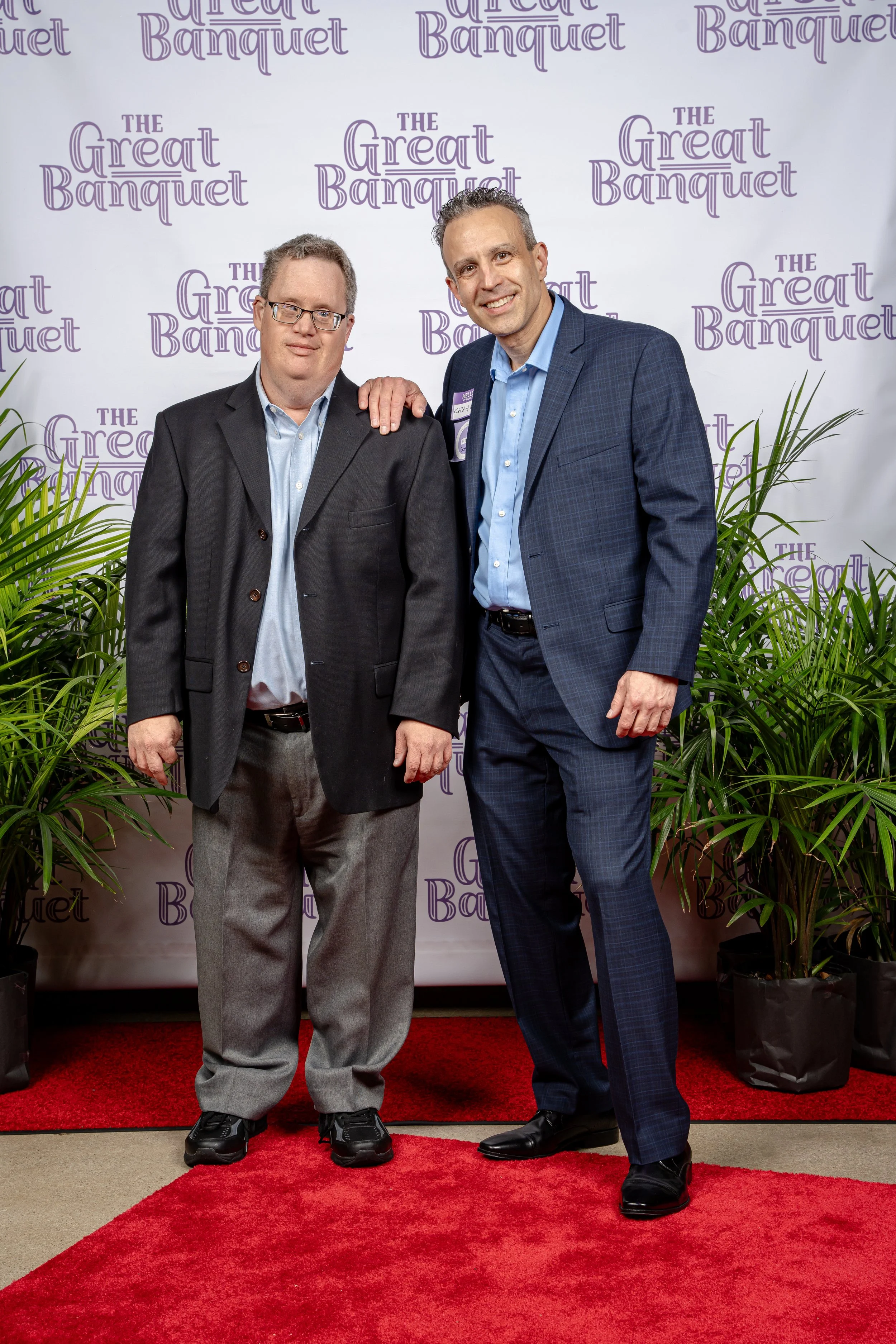 Two men standing on a red carpet in front of a backdrop that reads 'The Great Banquet'. One man has his arm around the other. There are large green plants on either side of them.