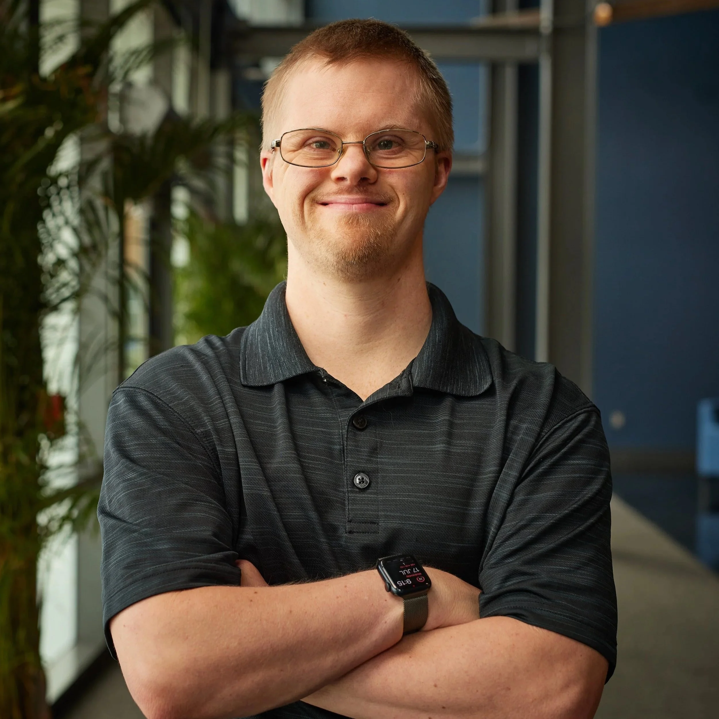 A young man with glasses, light brown hair, and a light beard, smiling with arms crossed, wearing a black polo shirt and a smartwatch, standing indoors with plants and office decor in the background.