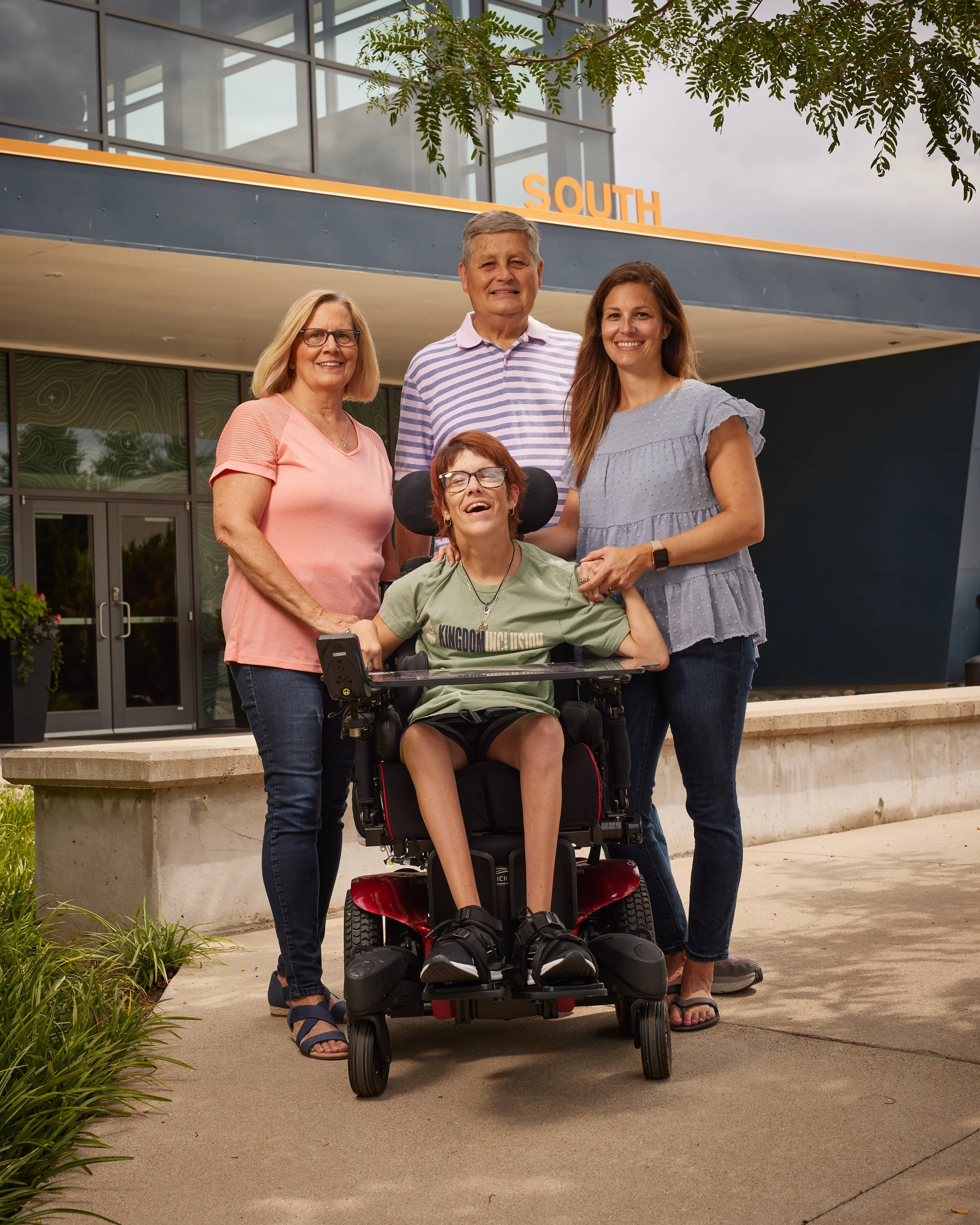 Group of five people outdoors in front of a building with the word South on it, including a woman in a wheelchair and four standing adults smiling.