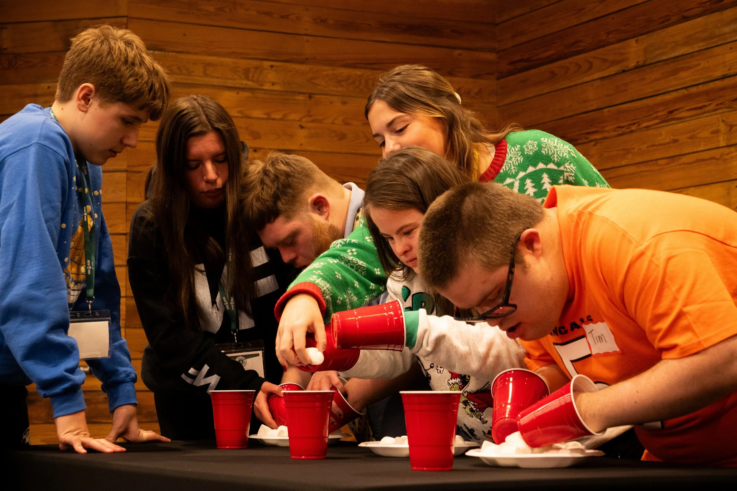 Group of people playing a game of cake relay race at a holiday party in a room with wood-paneled walls.