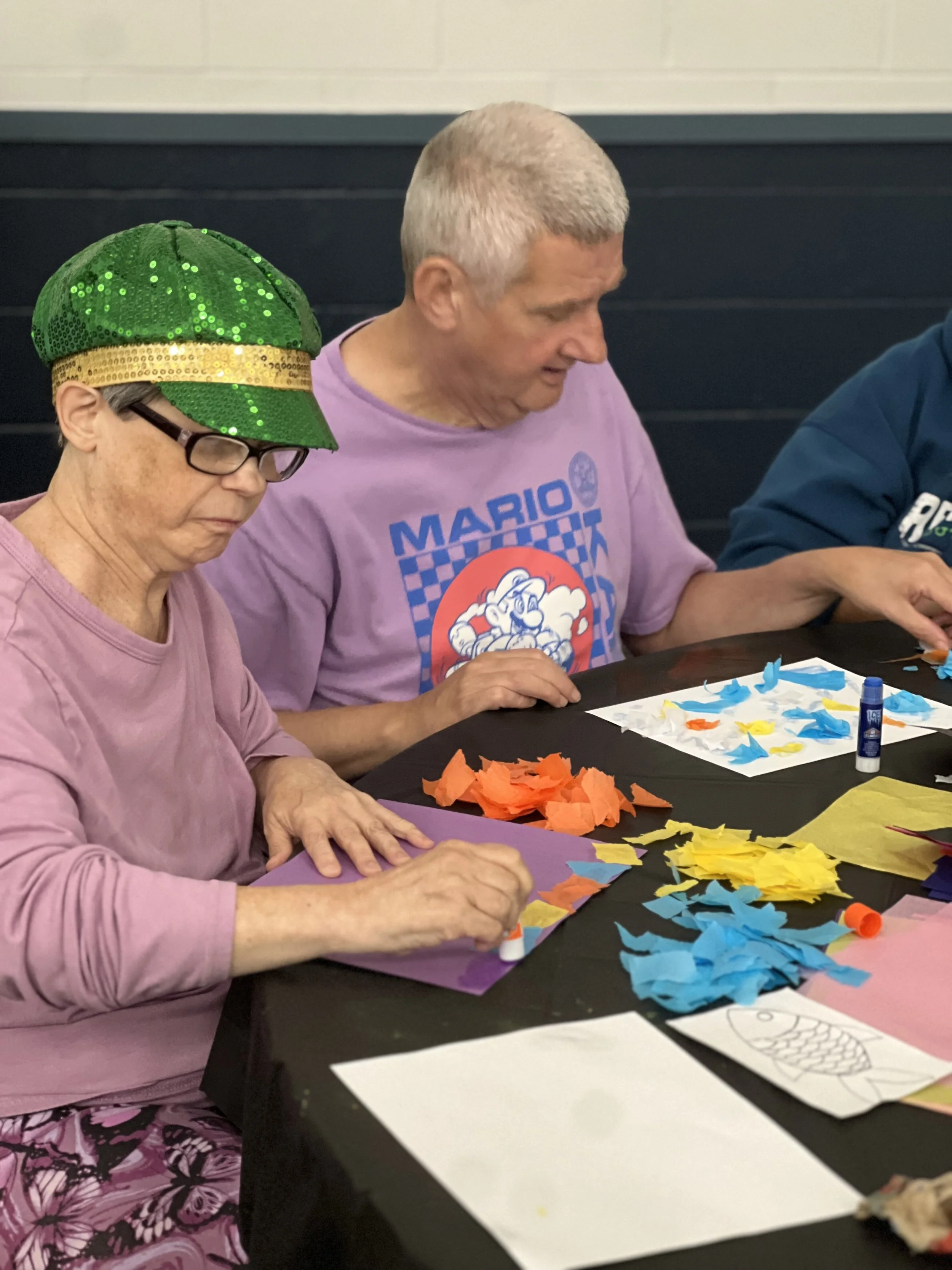 Two women seated at a table engaging in arts and crafts, with colorful tissue paper and glue sticks, one woman wearing a green and gold sequined cap and glasses, the other wearing a purple Mario T-shirt.