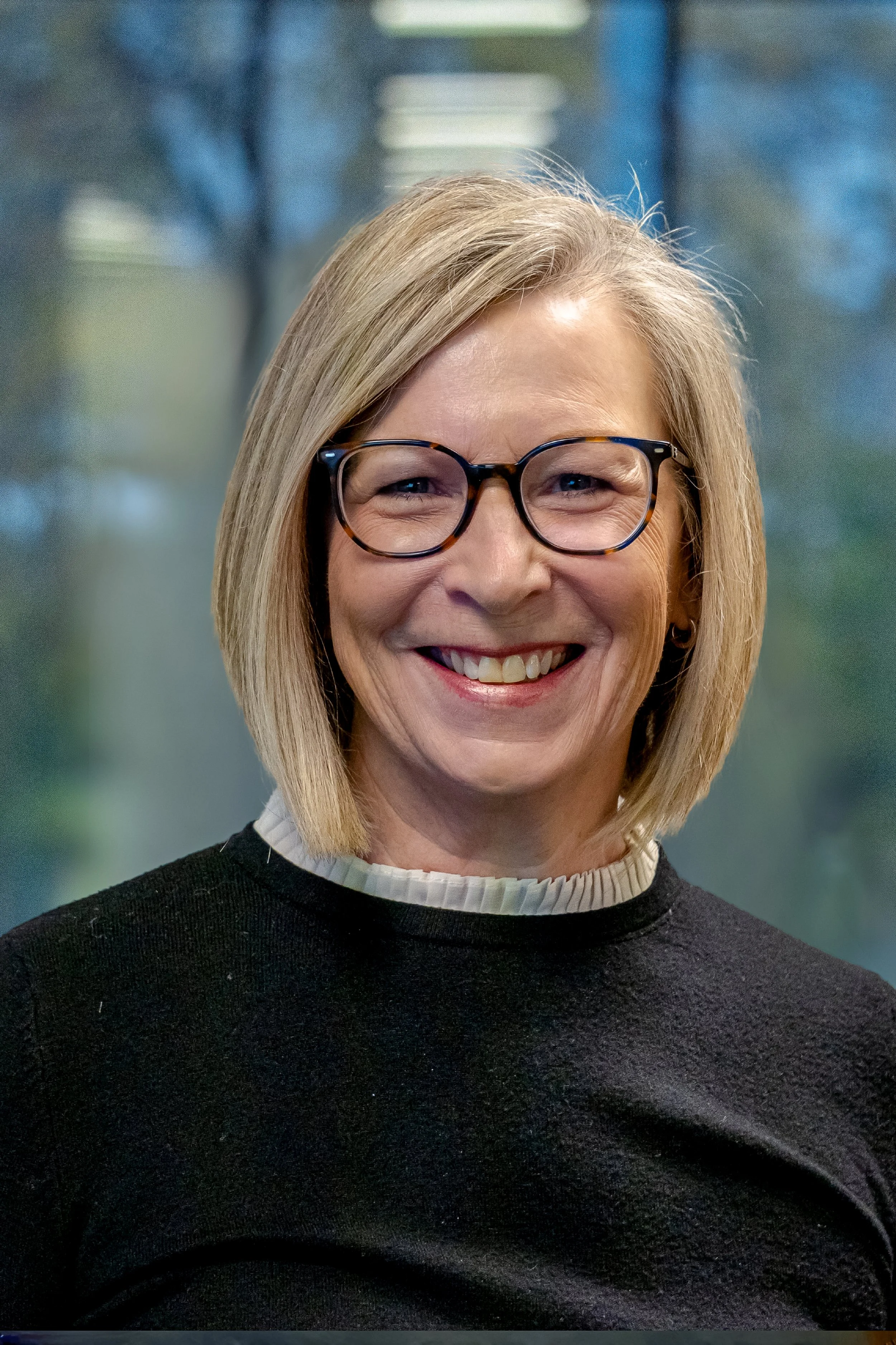 Portrait of a smiling middle-aged woman with blonde hair and glasses, wearing a black top with a white collar, standing in front of a blurred background of trees and sky.