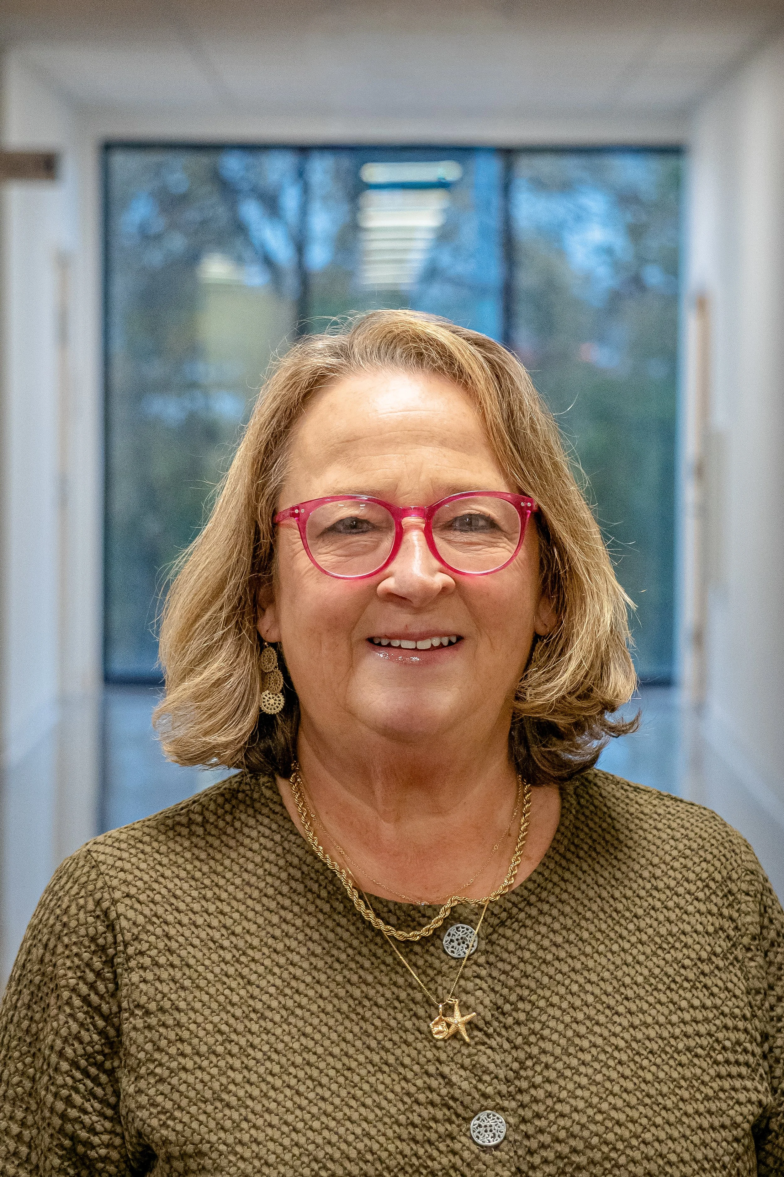 A middle-aged woman with shoulder-length blonde hair and red glasses smiling in a hallway with a window in the background.