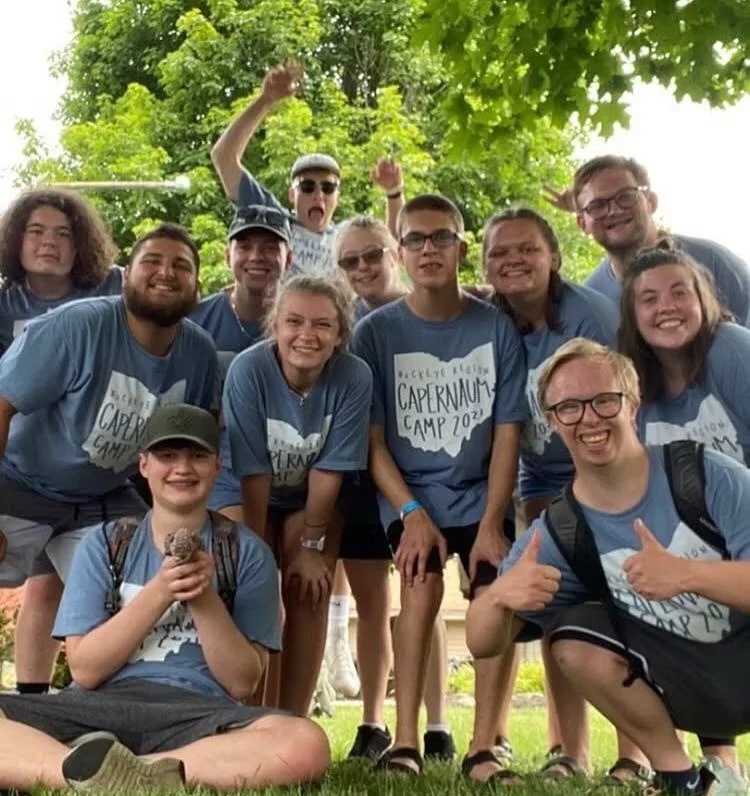 Group of smiling young people wearing matching blue T-shirts with 'Cappernaum Camp 2023' logo, posing outdoors with green trees in the background, some giving thumbs up or raising fists, one person holding a rock, most wearing glasses, showing excitement and camaraderie.